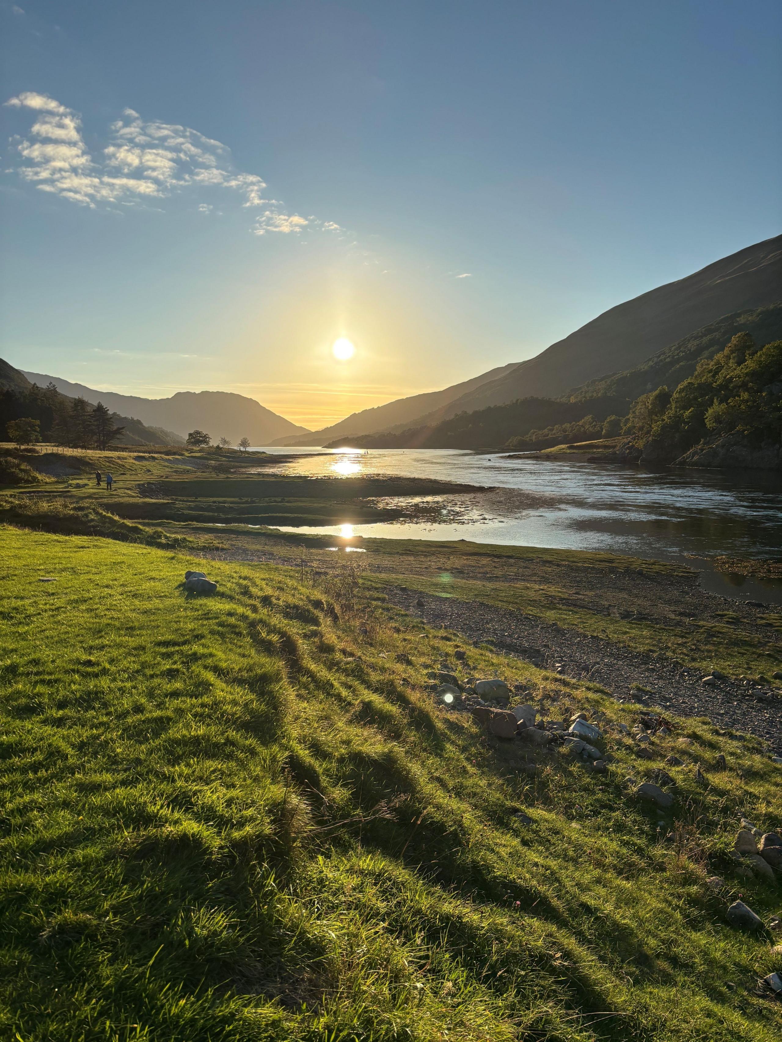 A vertical view of a loch snaking its way towards the sunset , hills in shadow all around and rich green grass in the foreground.