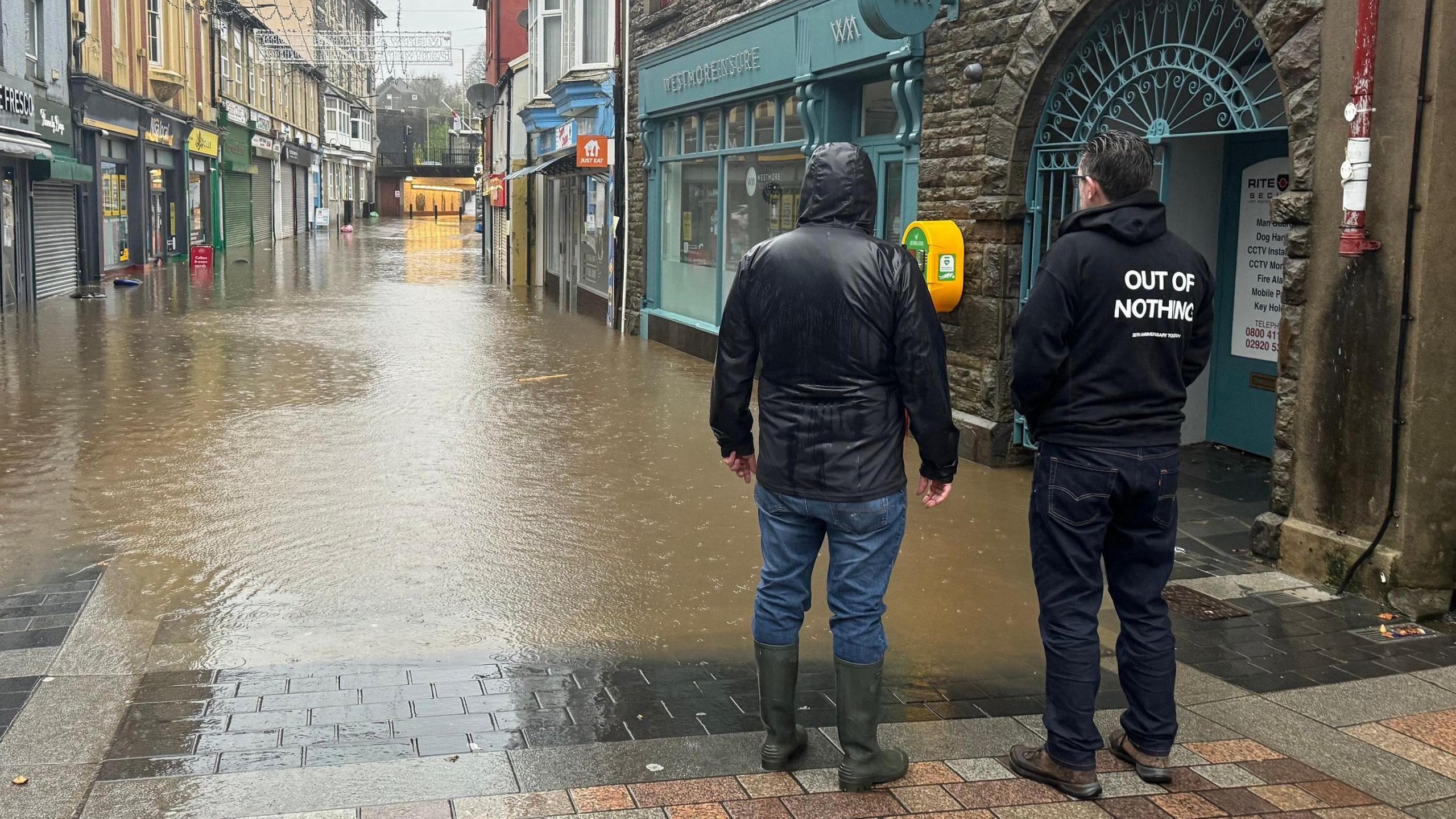 Two men stand in coats looking at flood waters in the high street of a town centre.