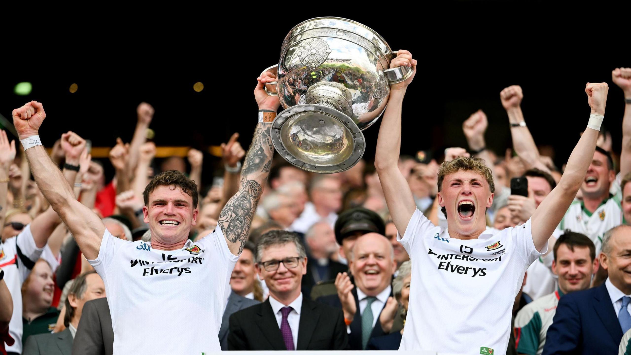 Two young men are standing with their backs to a large crowd in a stadium. They are both wearing white football tops and are holding the large, silver Sam Maguire trophy up between them. They are shouting and celebrating. 