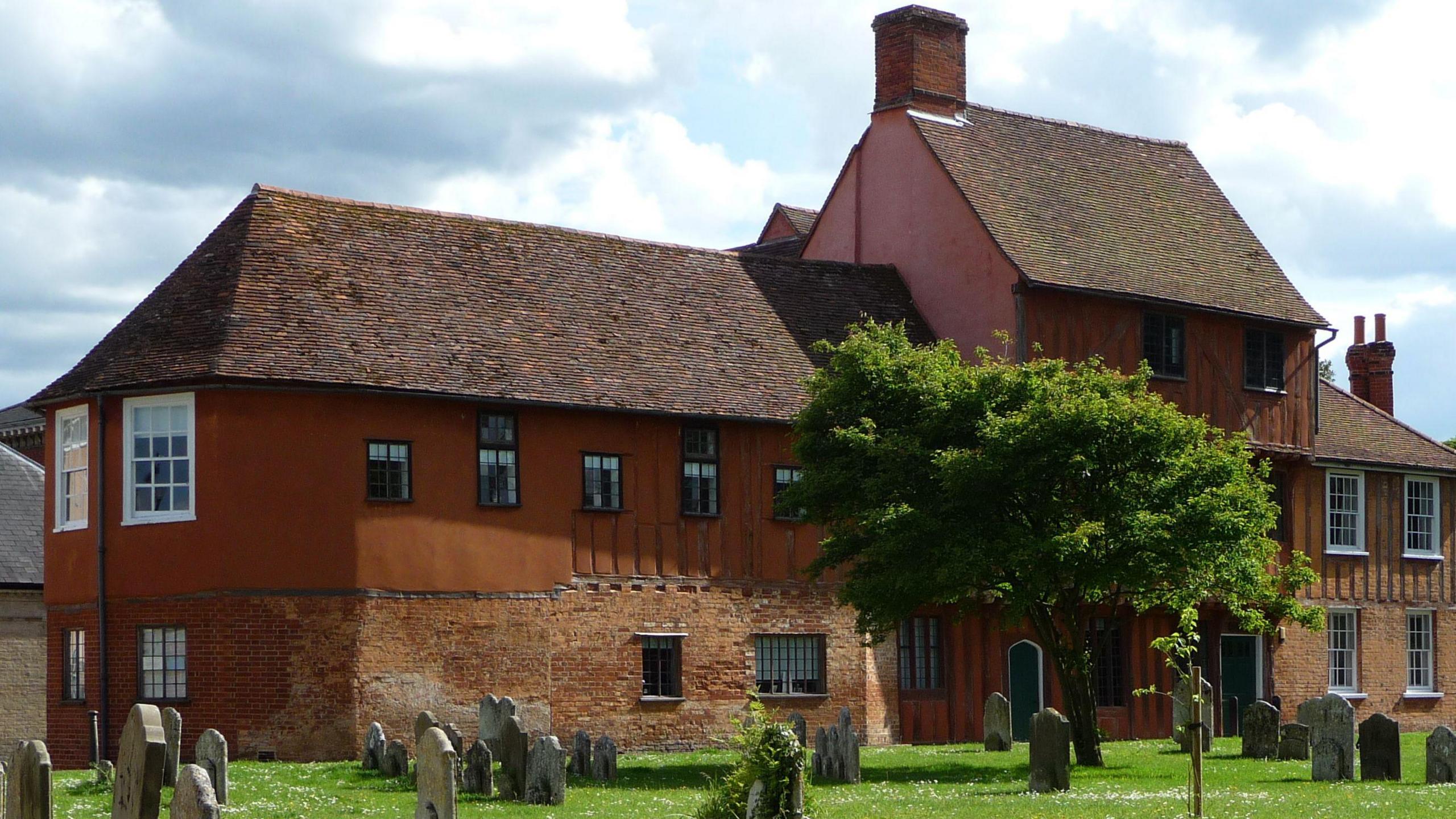 Hadleigh Guildhall is pictured during a sunny day. The building sits over two storeys with one middle part reaching up to three storeys. There are several gravestones outside the building along with a tree.