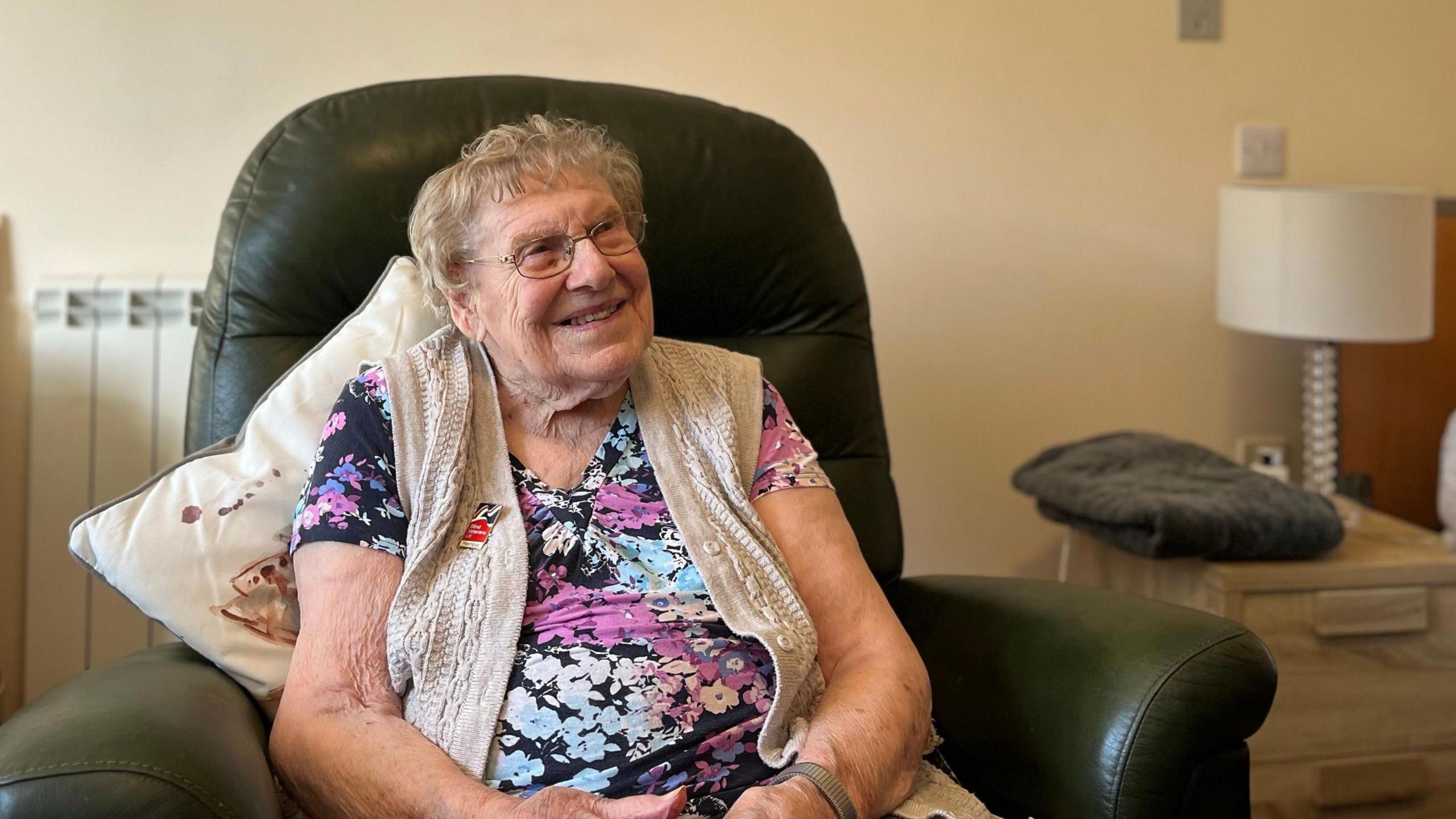 A smiling woman with glasses in a flowery dress and a pale sleeveless cardigan sitting on a green  chair with a cushion behind her.