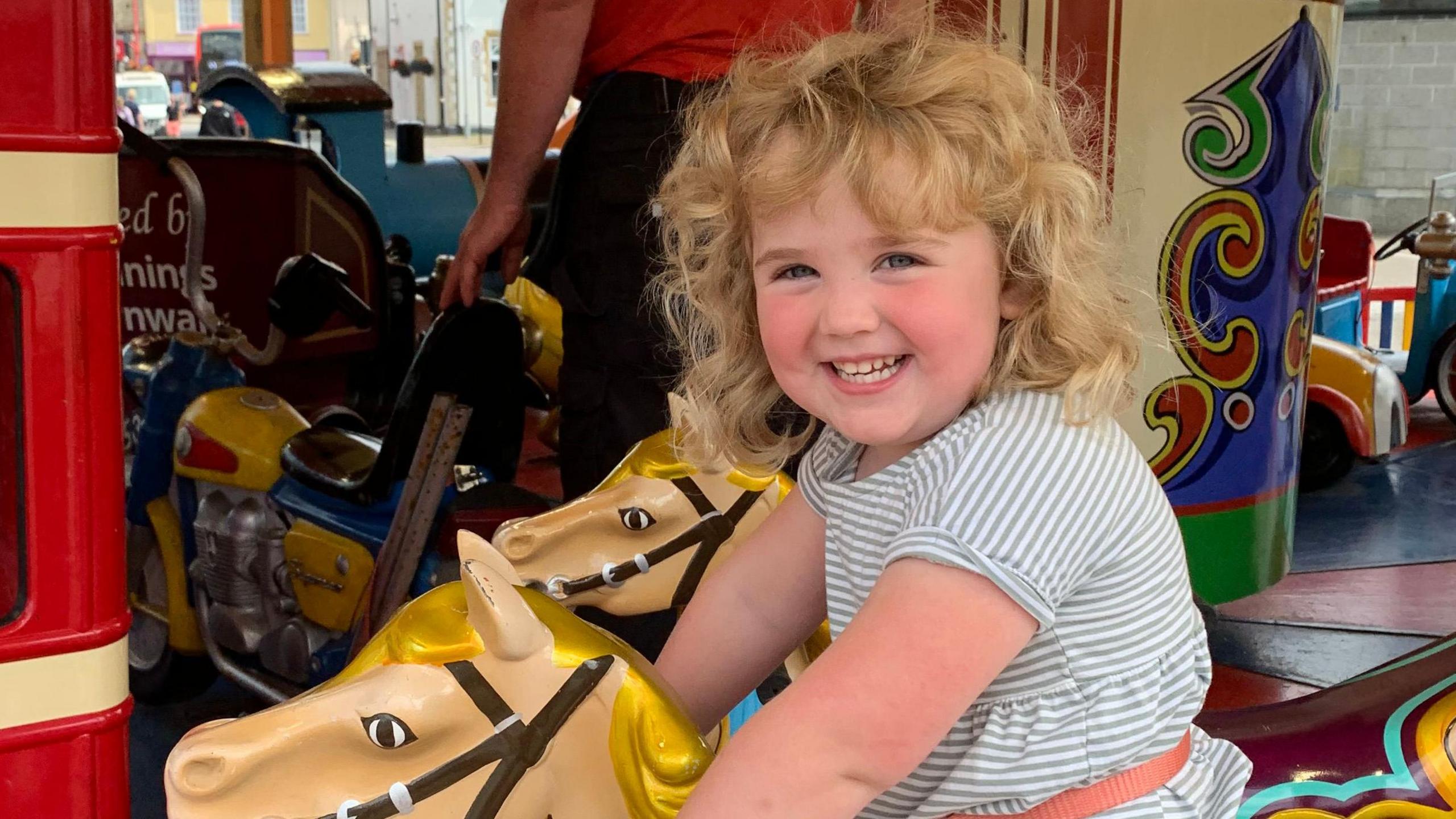 A little girl in a white and blue dress is smiling on a carousel. 