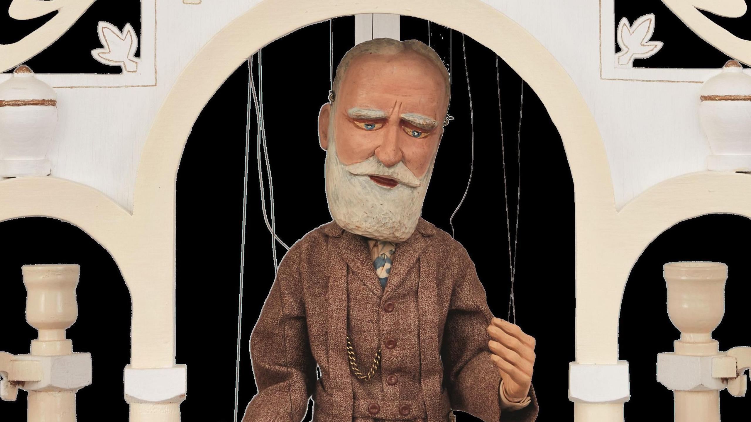 Puppet of a bearded older man in a suit. The puppet, dressed in tweeds, is under an ornate archway.