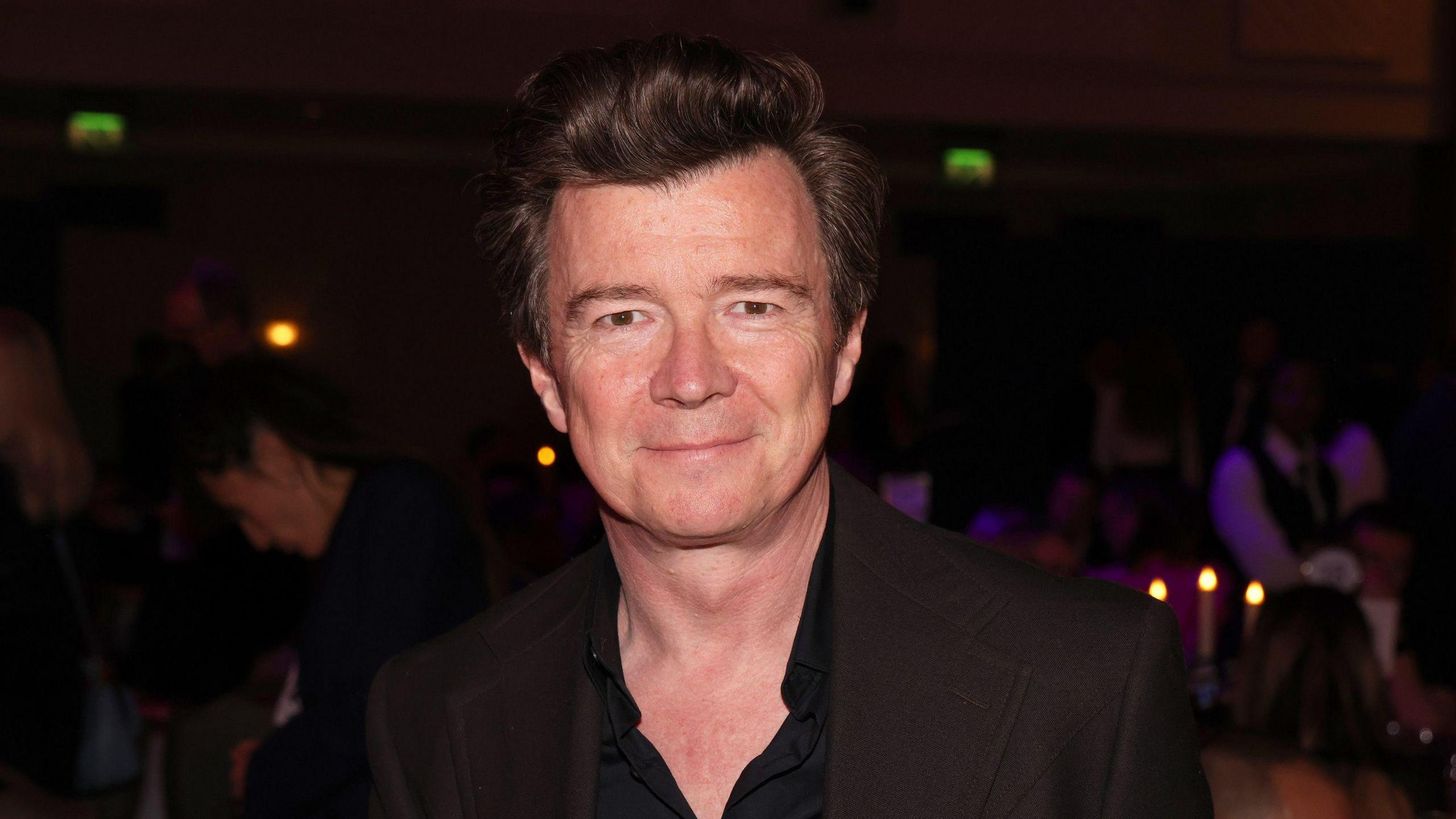 Rick Astley smiles at the camera. He is wearing a black shirt, unbuttoned to his chest, and is standing in a dark room at an awards ceremony