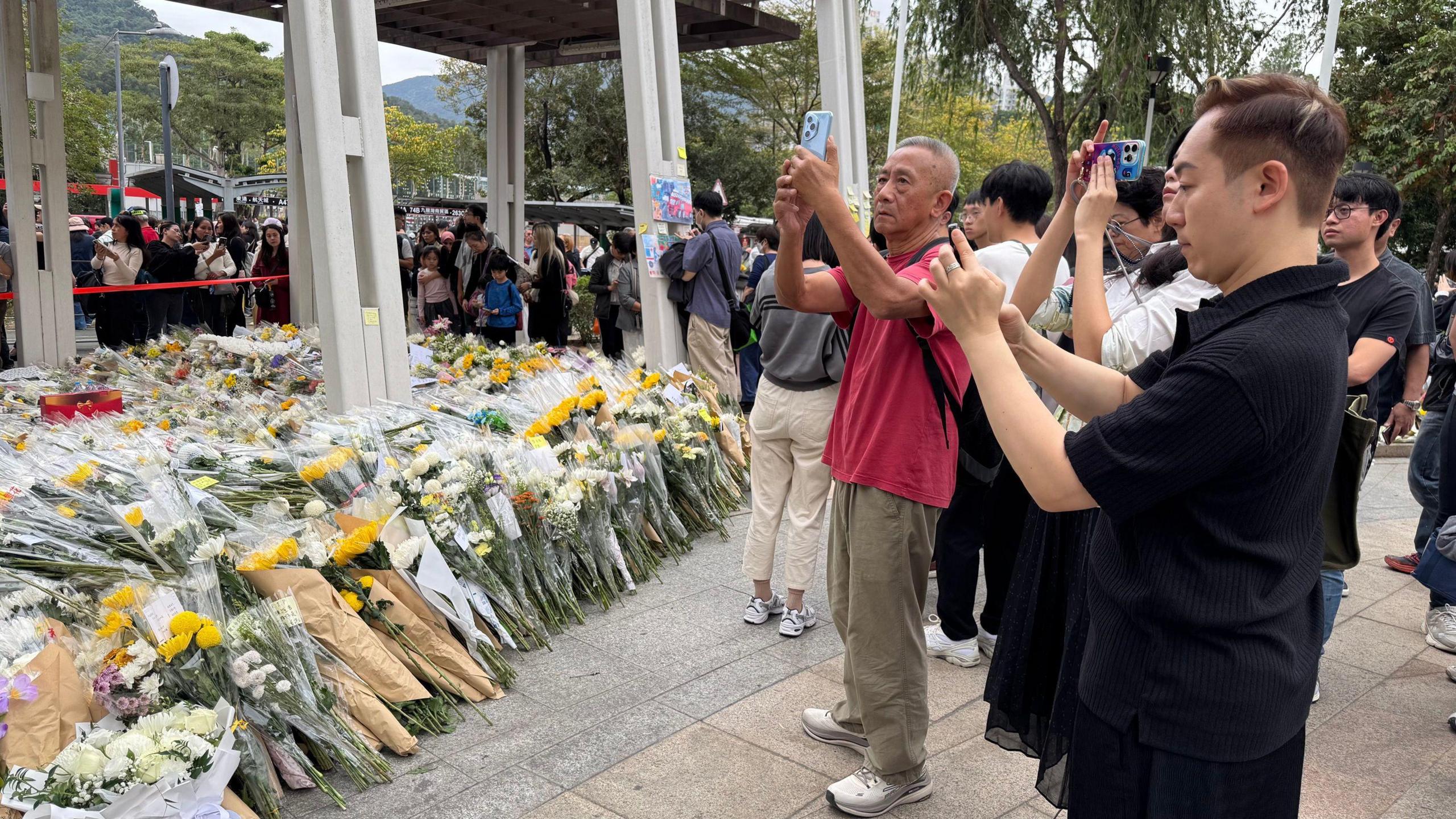 Hundreds of flower bouquets on the left. On the right are people who are queuing, some of whom are taking photos