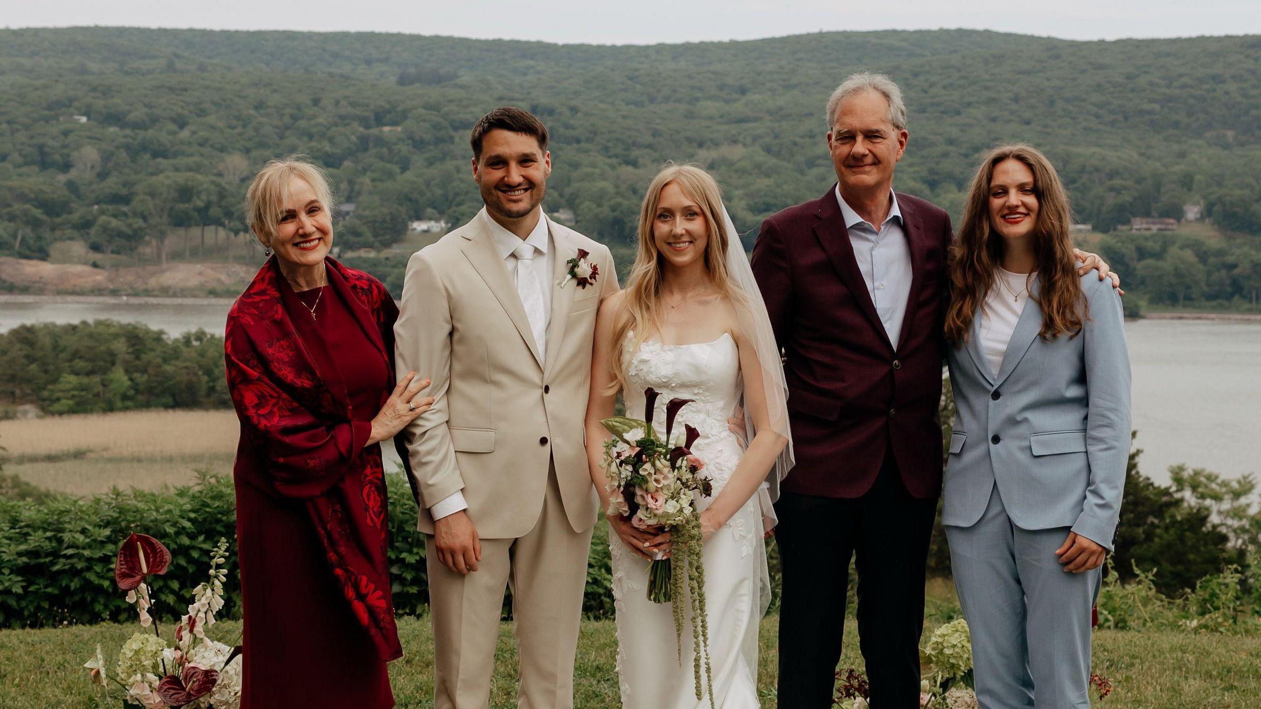 Michele Heisler stands next to her son on his wedding day, flanked by her husband and daughter. She wears a red velvet shawl and dress, while her son wears a beige suit.