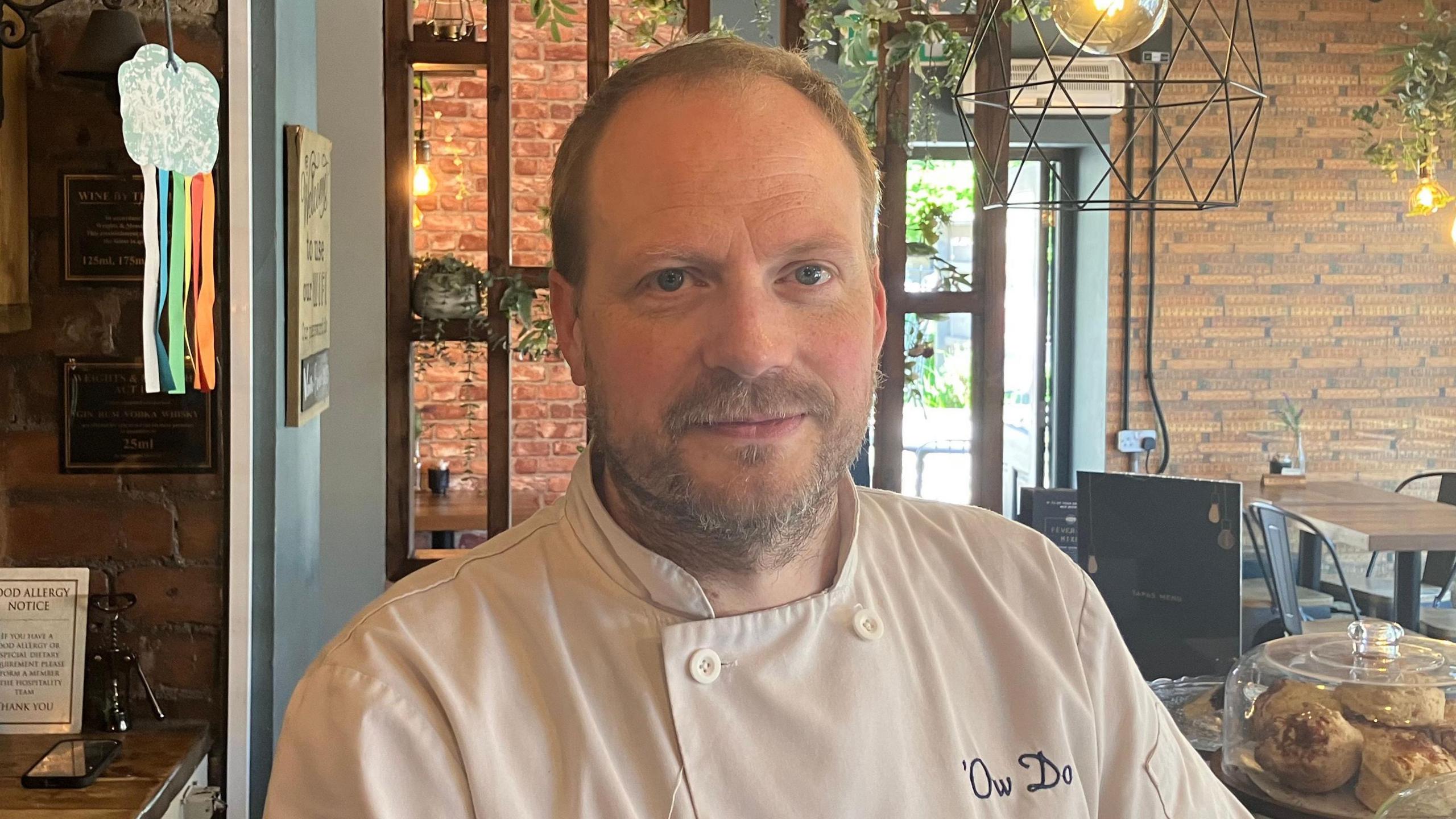 A man with short light brown hair stands in a cafe. He is wearing chef whites
