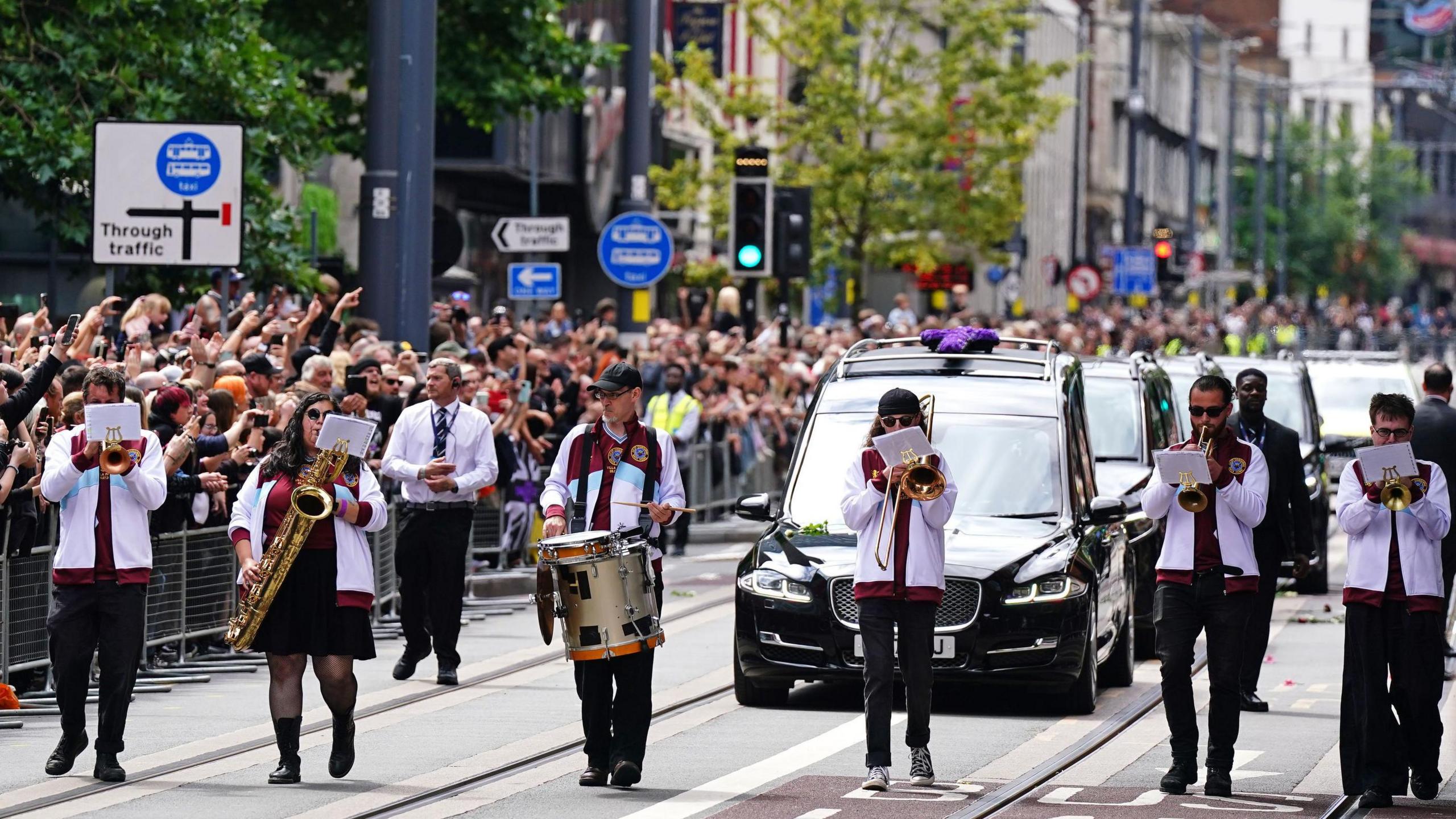 Six people wearing white jackets with red trim are walking down the middle of a road, each playing a musical instrument. There are large black funeral cars behind and crowds of people lining the street of a main high street.