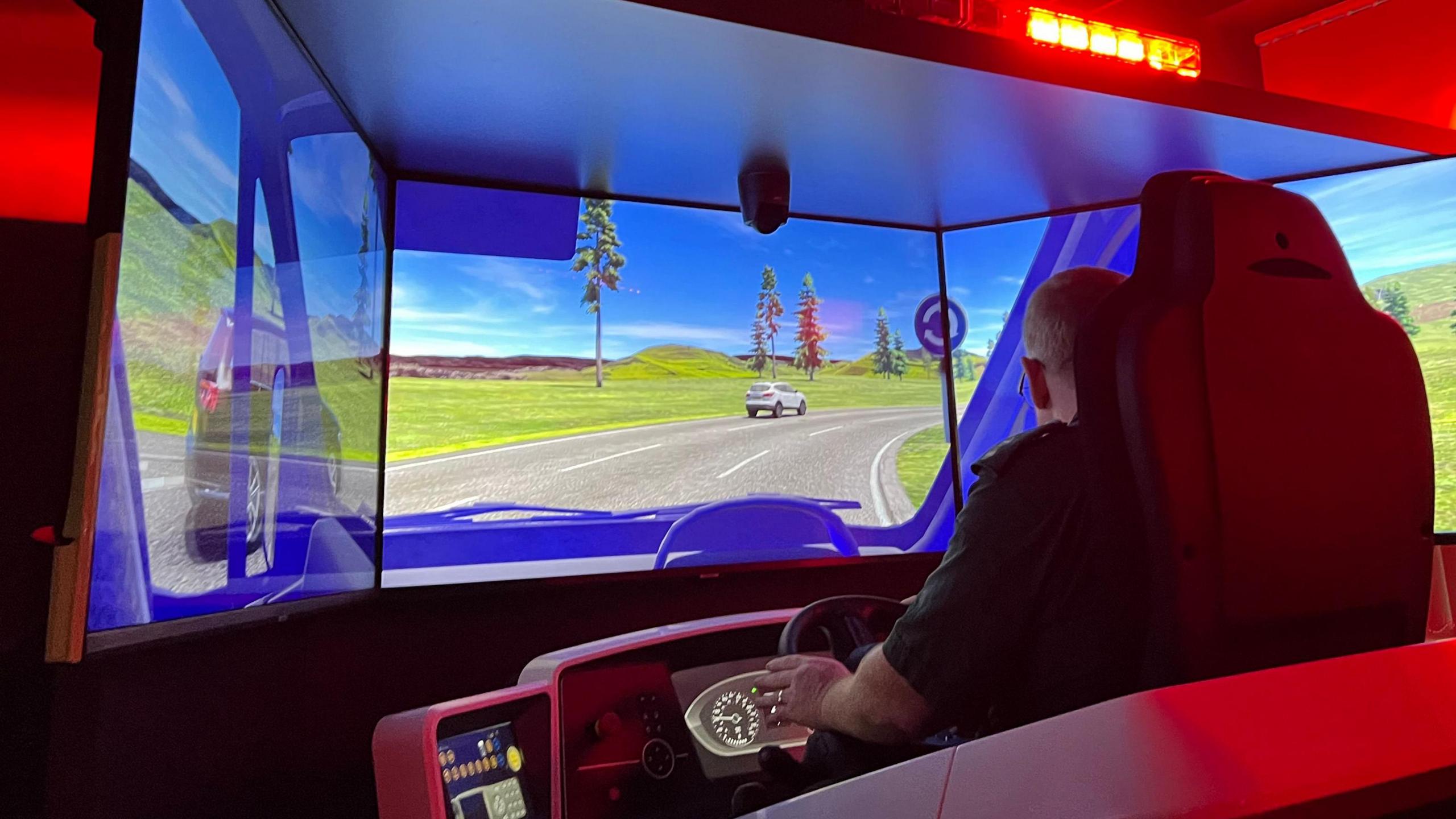 A man sitting in front of a driving simulator. A red flashing light above his screen is casting a red glow and computer screens in front of him mimic an ambulance windscreen. On the computer screen a road can be seen, with vehicles ahead and grass and trees on the verges.