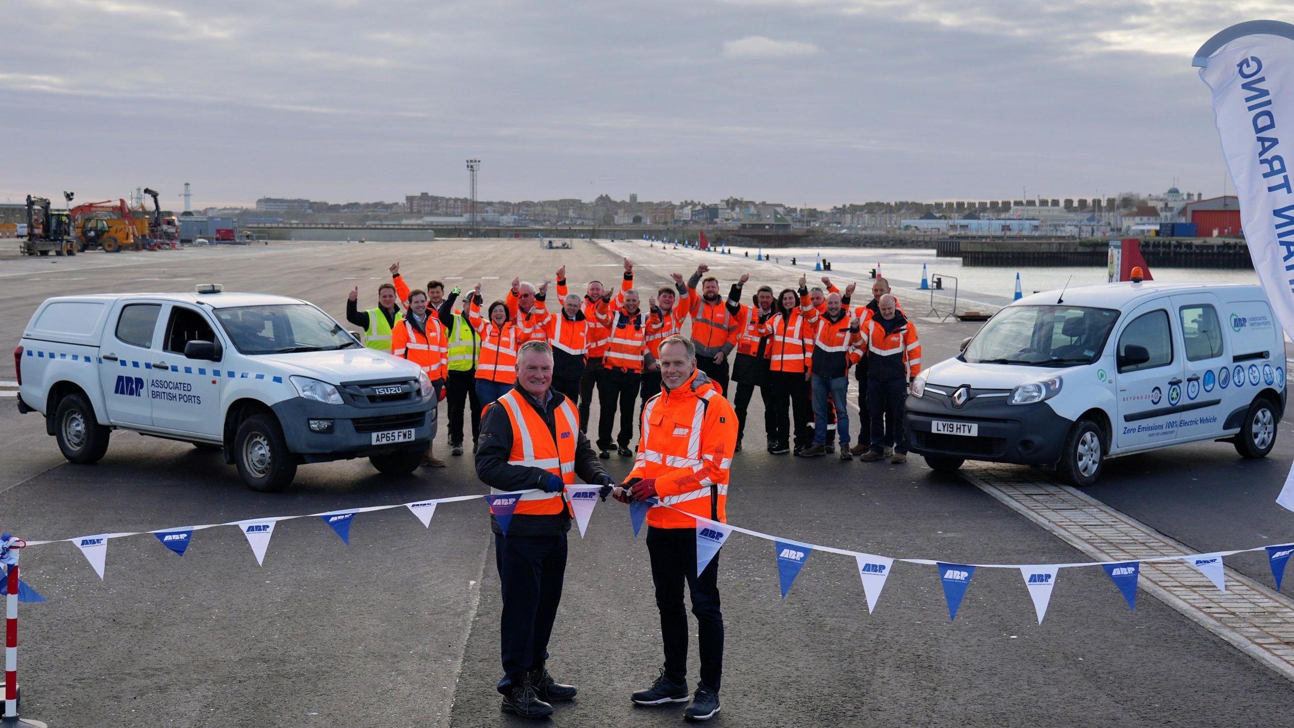 Two people in orange fluorescent jackets cut a ribbon decorated with blue and white flags, with two vans and group of cheering people in the background