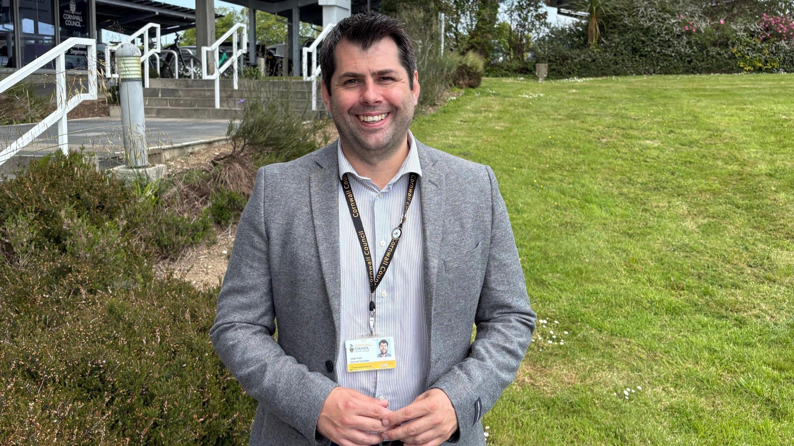 Leigh Frost is smiling as he stands in a garden with County Hall behind him. He has dark hair and brown eyes and is wearing a grey jacket and a grey and white striped shirt and a lanyard with his Cornwall Council identification on it.