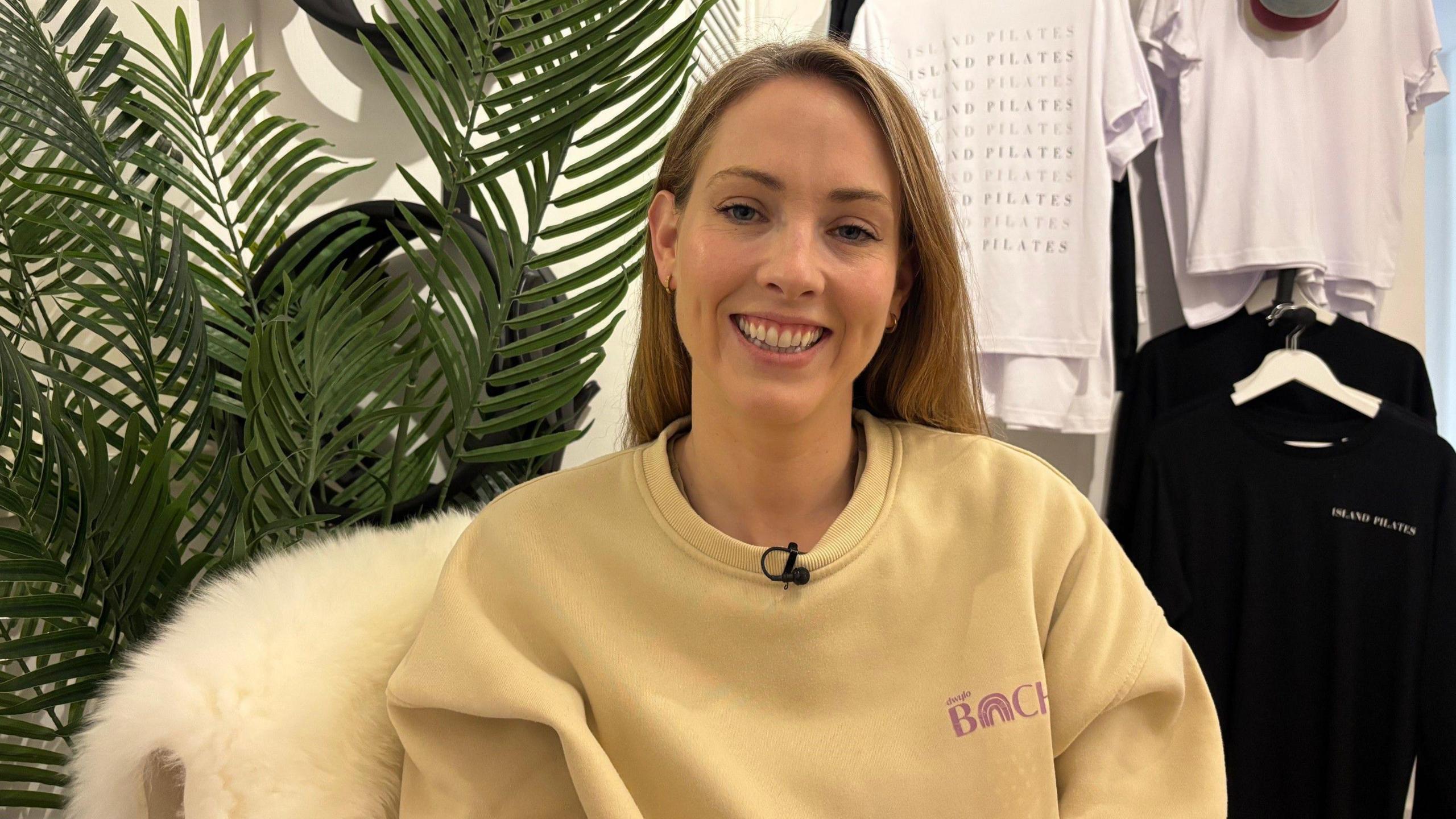 Catrin hughes sits on a white fur chair, wearing a yellow sweatshirt. She has light blonde/ brown hair. 