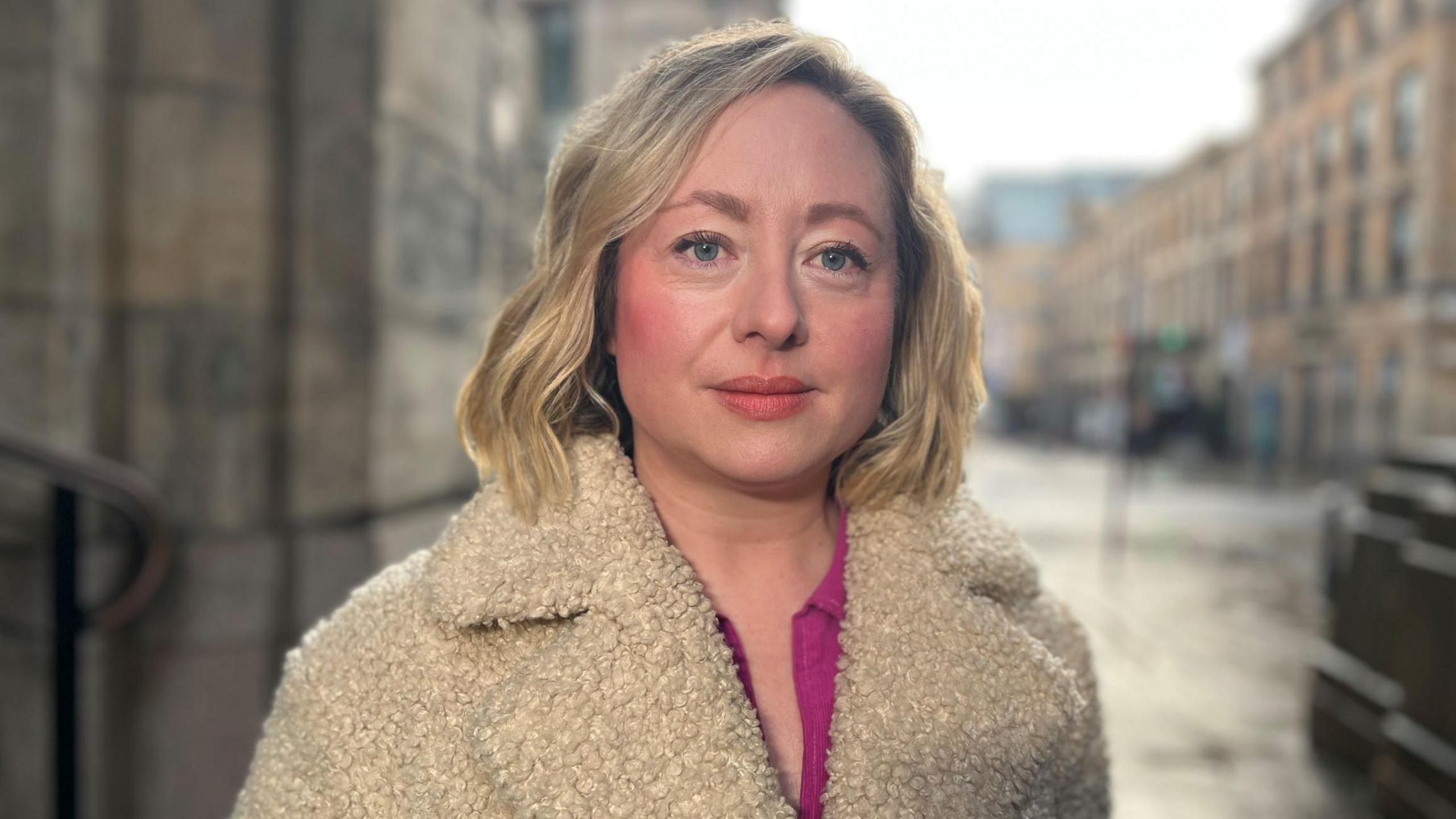 Catriona Stewart looking into the camera while standing outside in Glasgow City Centre. She has blonde hair, a pink top and a winter coat on. 