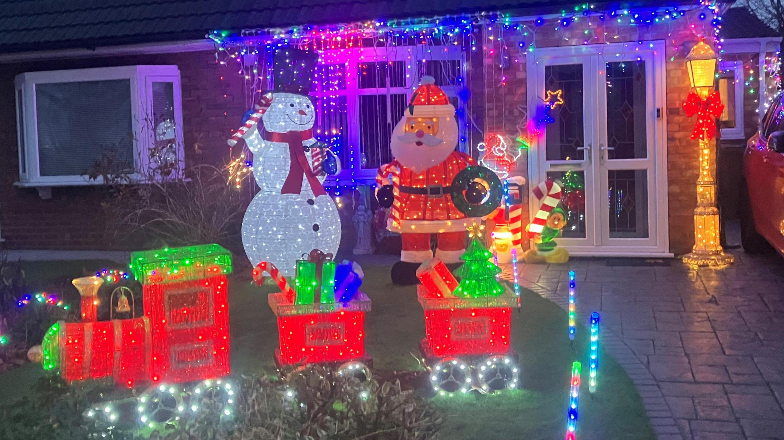 A lawn in front of a property covered in Christmas lights and decorations, including a train filled with presents, a large snowman holding a candy cane, a Father Christmas figure and lots of other Christmas adornments. 