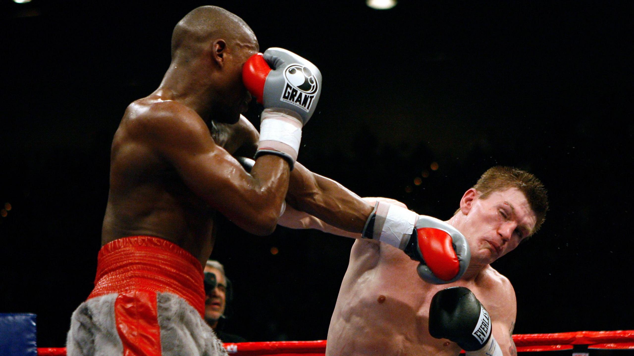 Floyd Mayweather Jr landing a left hook on Ricky Hatton. Mayweather is wearing grey shorts with a red trim. Hatton is wearing black gloves.