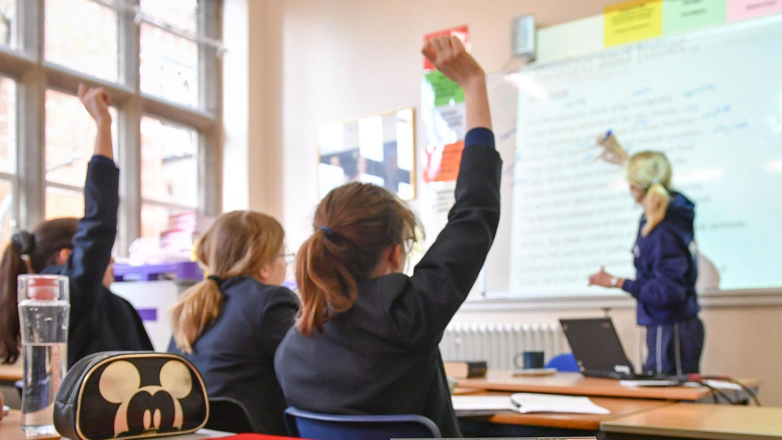 A female teacher writing on a whiteboard in a classroom, and three girls with their back to the camera, wearing dark blazers. The first and third girl have their hands raised. 