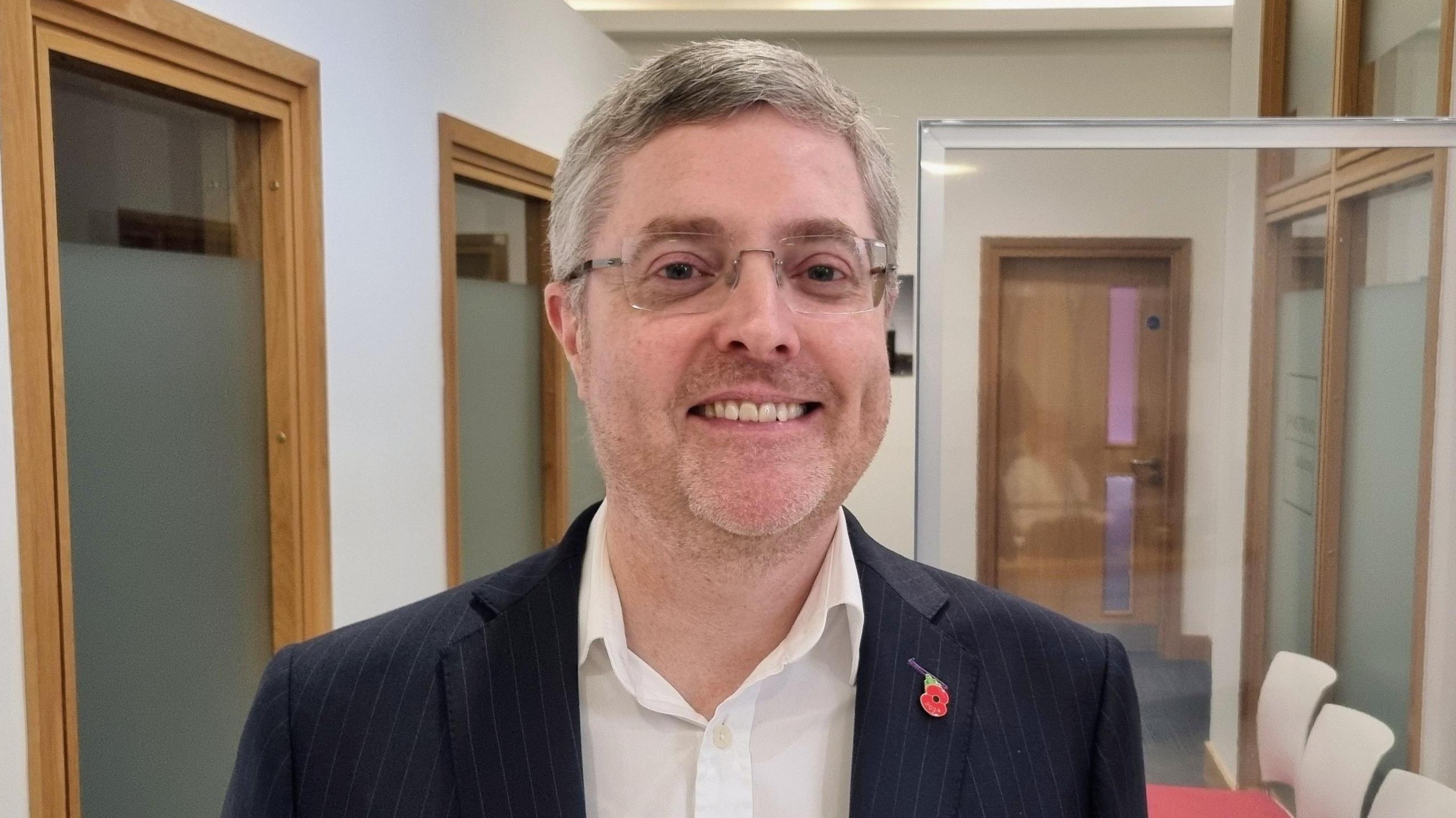 Philip Armstrong in black blazer and white shirt, smiling in a corridor with doors and chairs in background