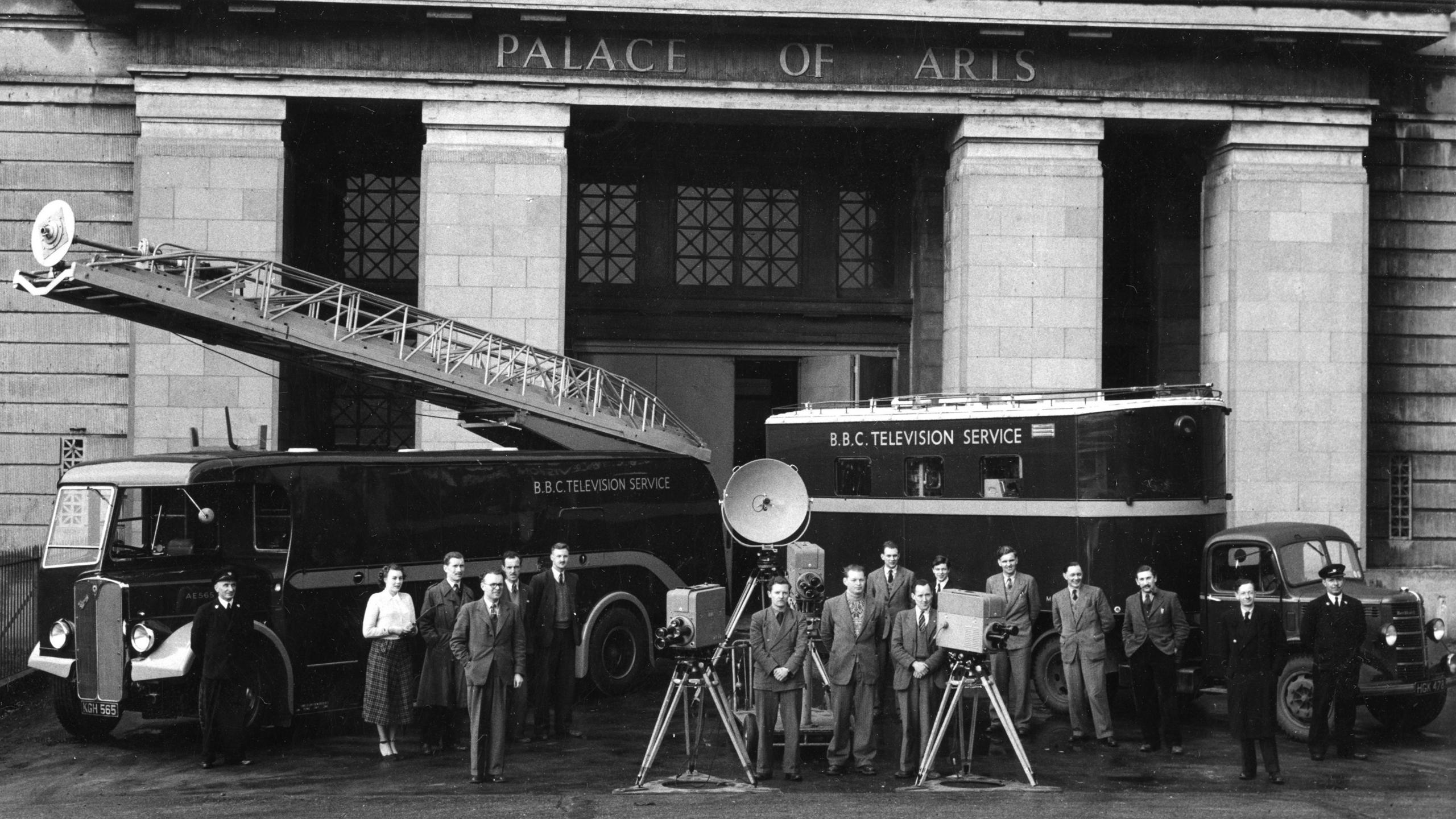 A black and white posed photograph of 16 people and two trucks in front of an ornate stone building with Palace of Arts written across the grand-looking entranceway. Both trucks are labelled 'BBC Television Service', and the one on the left has a large crane mounted on the top. The people are standing in a row in front of the trucks, along with three television cameras with turret lenses, mounted on tripod stands.