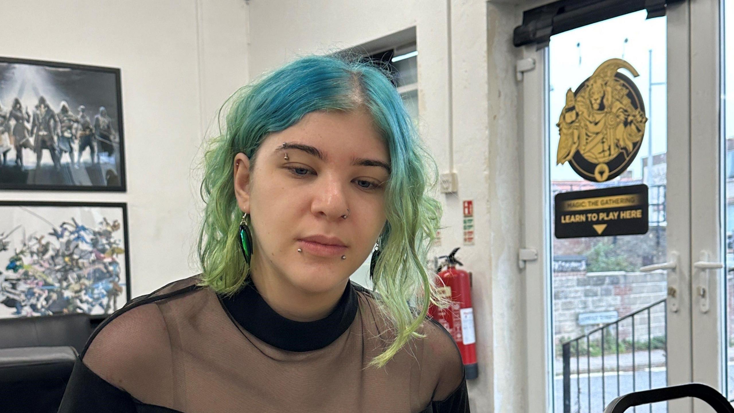 Evie is pictured looking away from the camera while she sits. She has blue and green hair which is cut to her shoulders. She has several piercings on her face as well as ear piercings. She is wearing a black top which is sheer on her shoulders.