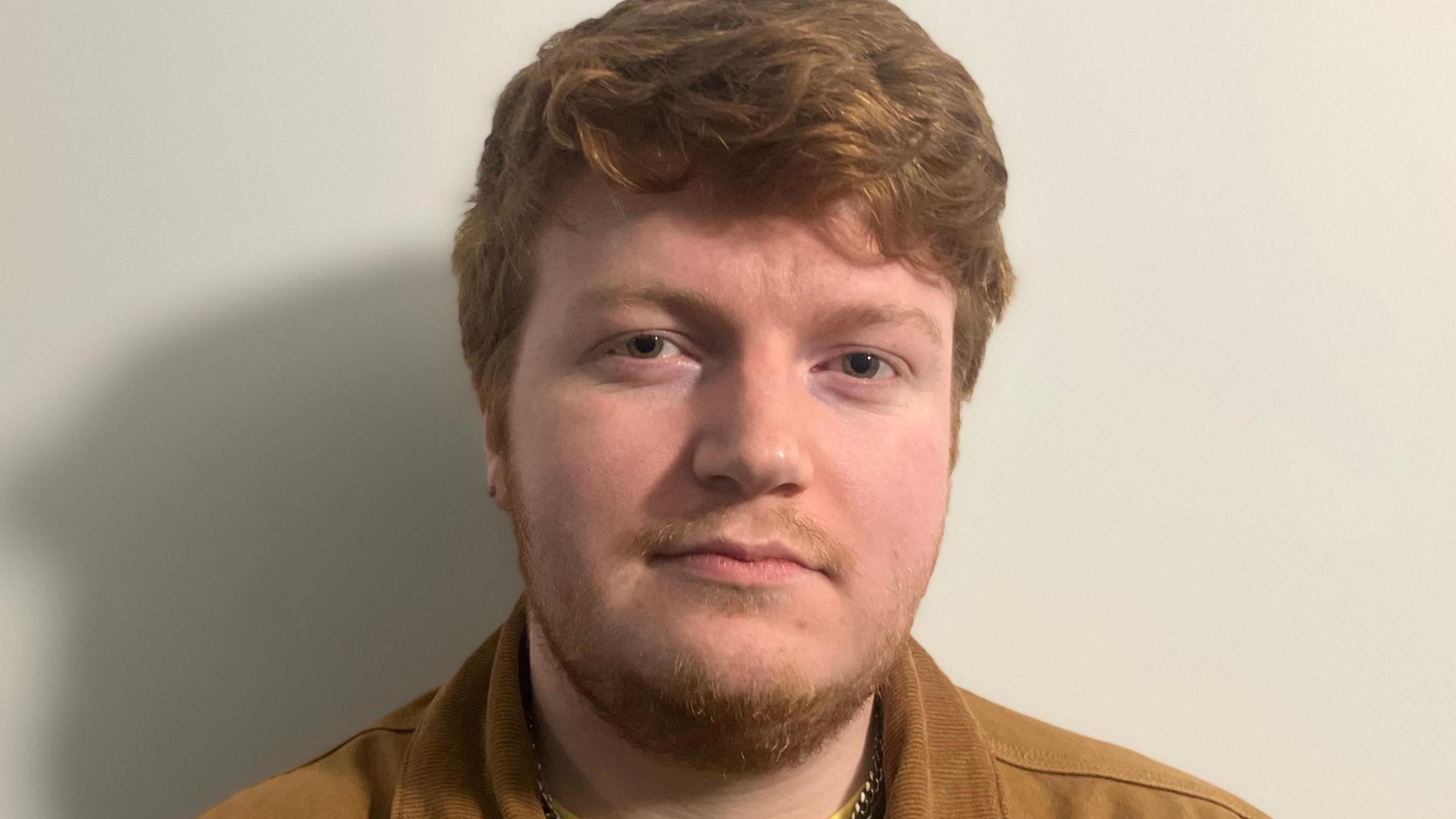 A man with ginger hair leans against a wall. He wears a brown shirt and a black top with a yellow seam.
