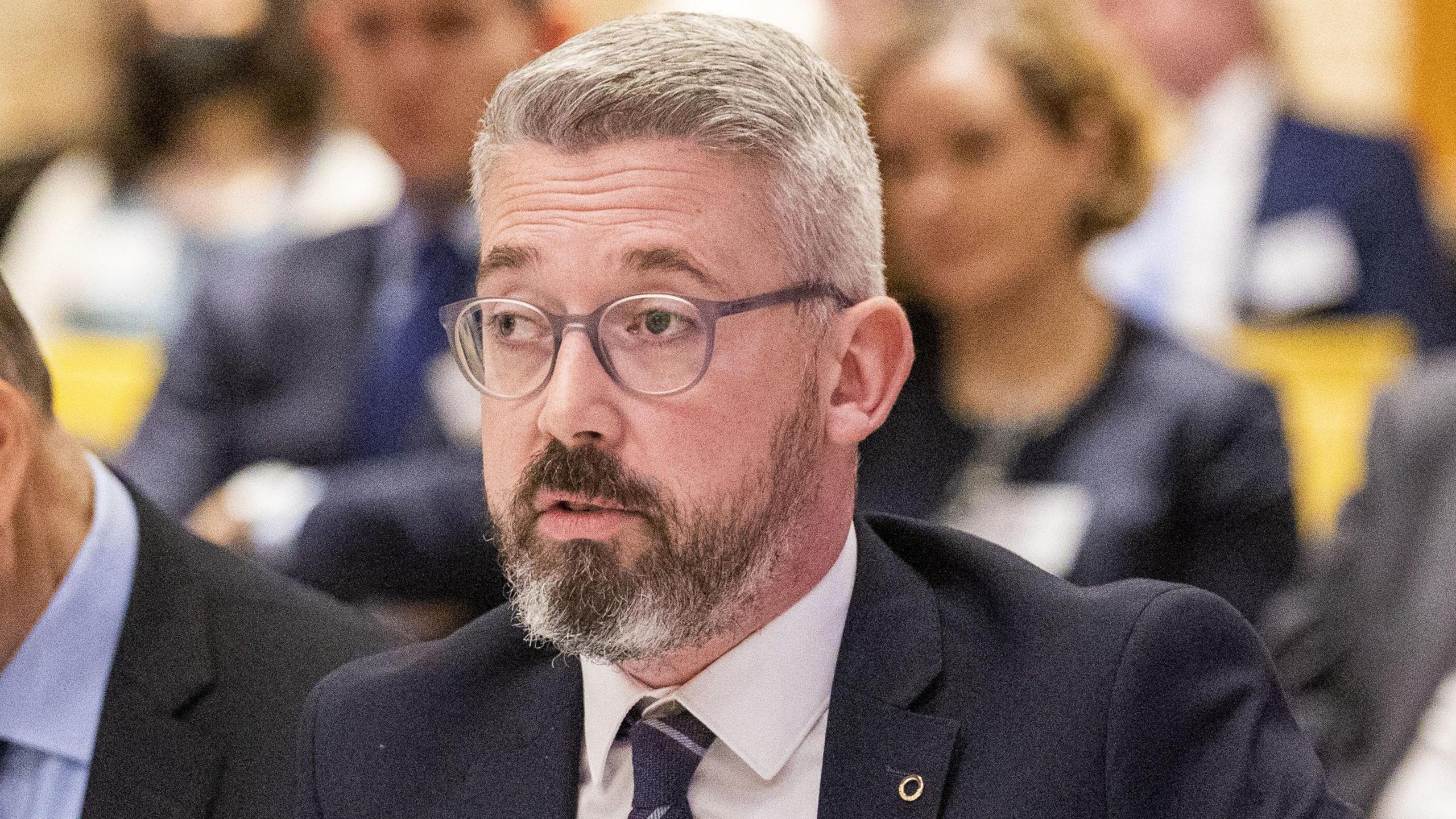 Niall Ó Donnghaile, pictured from the neck up, wearing a suit and white shirt. He has grey hair and is wearing glasses.