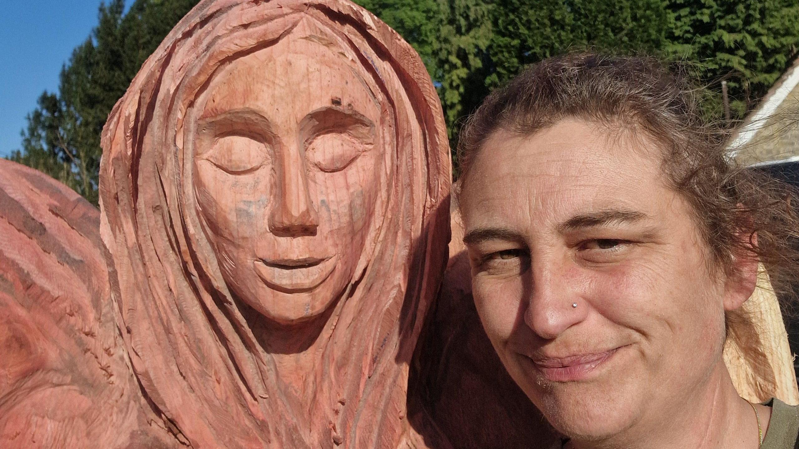 Lisa Langley has short brown hair pulled tightly over her head. She has a gold nose stud in her left nostril and a light green T-shirt on. She is standing right next to the carved angel's face, which is life-size.