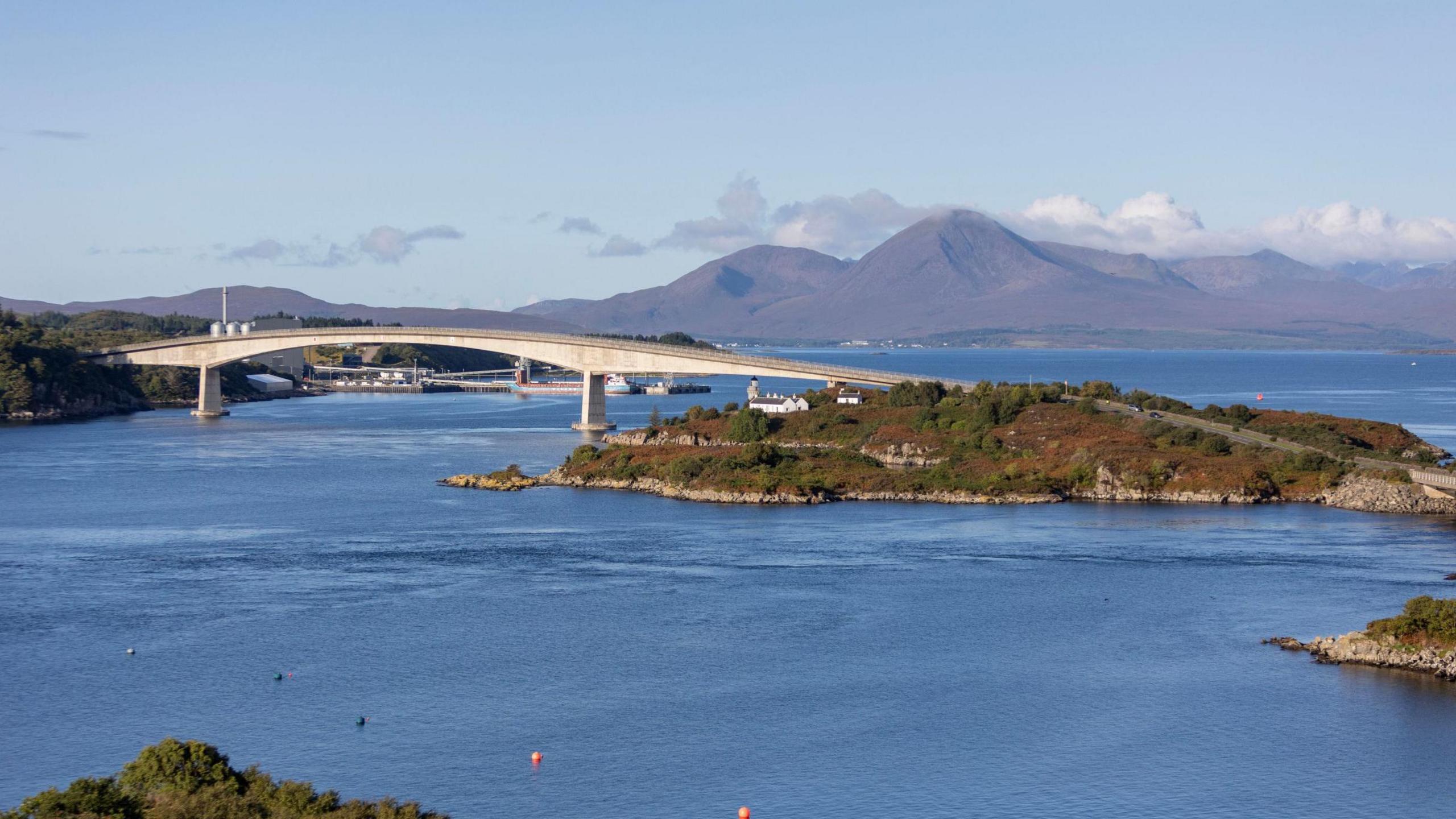 Skye Bridge pictured on a bright, sunny day. Behind the bridge is a range of hills, and the crossing spans calm, blue waters.