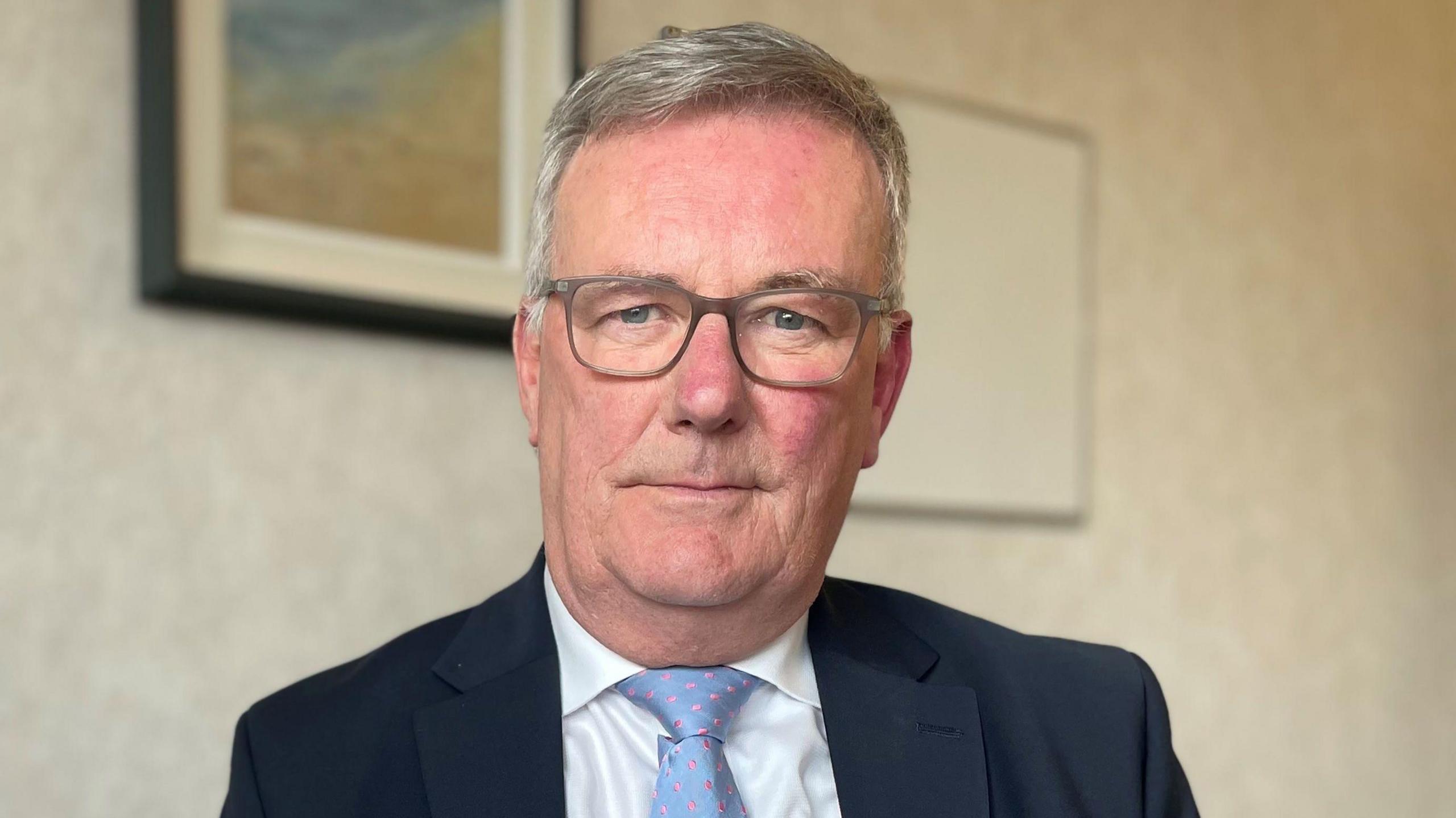 Mike Nesbitt looks at the camera. The health minister has short, grey hair and is wearing a pair of grey square-framed glasses, a suit jacket, a blue tie with pink spots and a white, collared shirt. He is sitting in a white room with some paintings behind him but they are blurred.