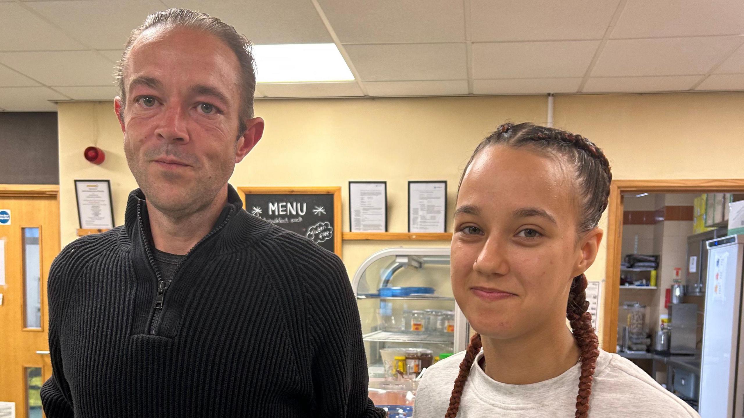 Service coordinator Wayne Twitchell and volunteer Larnie Perrett pictured in the cafe space. Mr Twitchell is wearing a dark coloured fleece and Ms Perrett (right) has her hair pulled back into two french braids and wears a grey sweatshirt. 