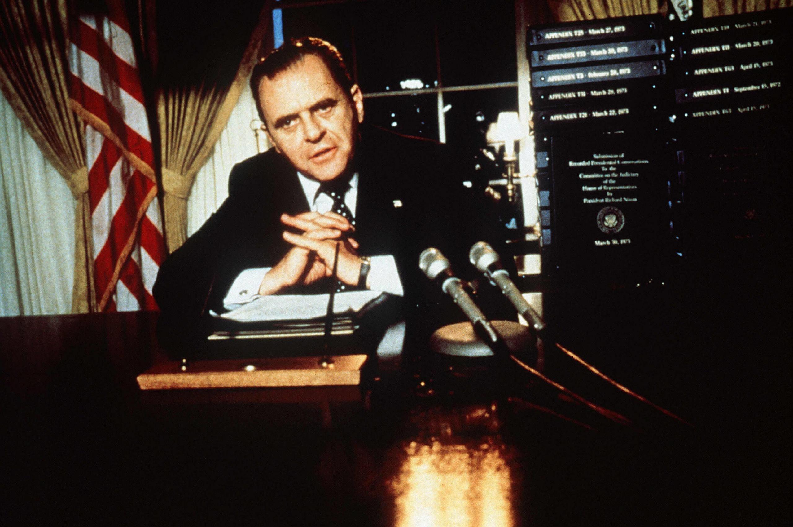 Hopkins as President Nixon, hair greased back his arms folded looking to camera with two microphones on his dark desk. US flag and curtains in background
