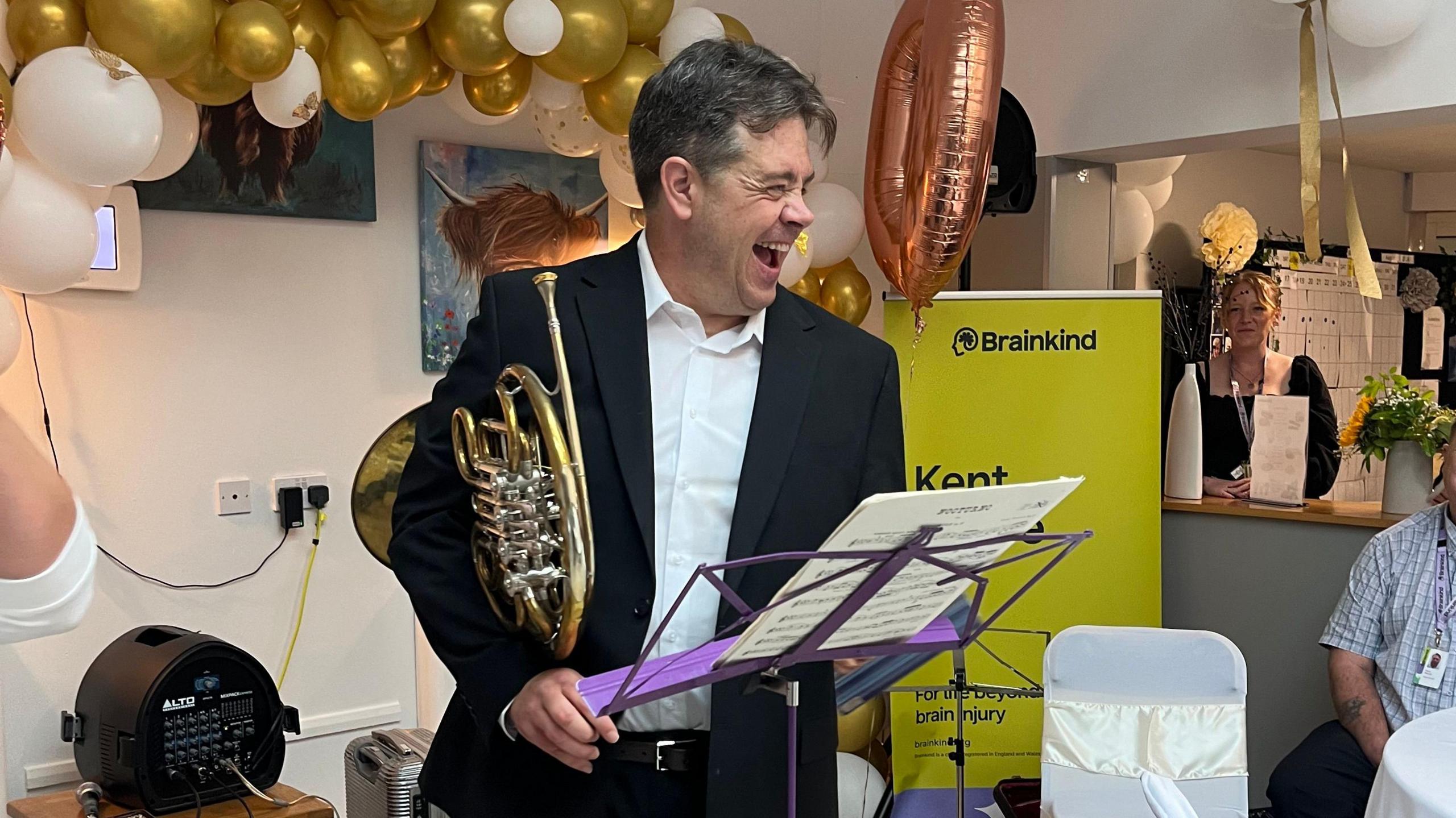 Rupert Johnston, smiling, holding a French Horn under his arm, at a party 