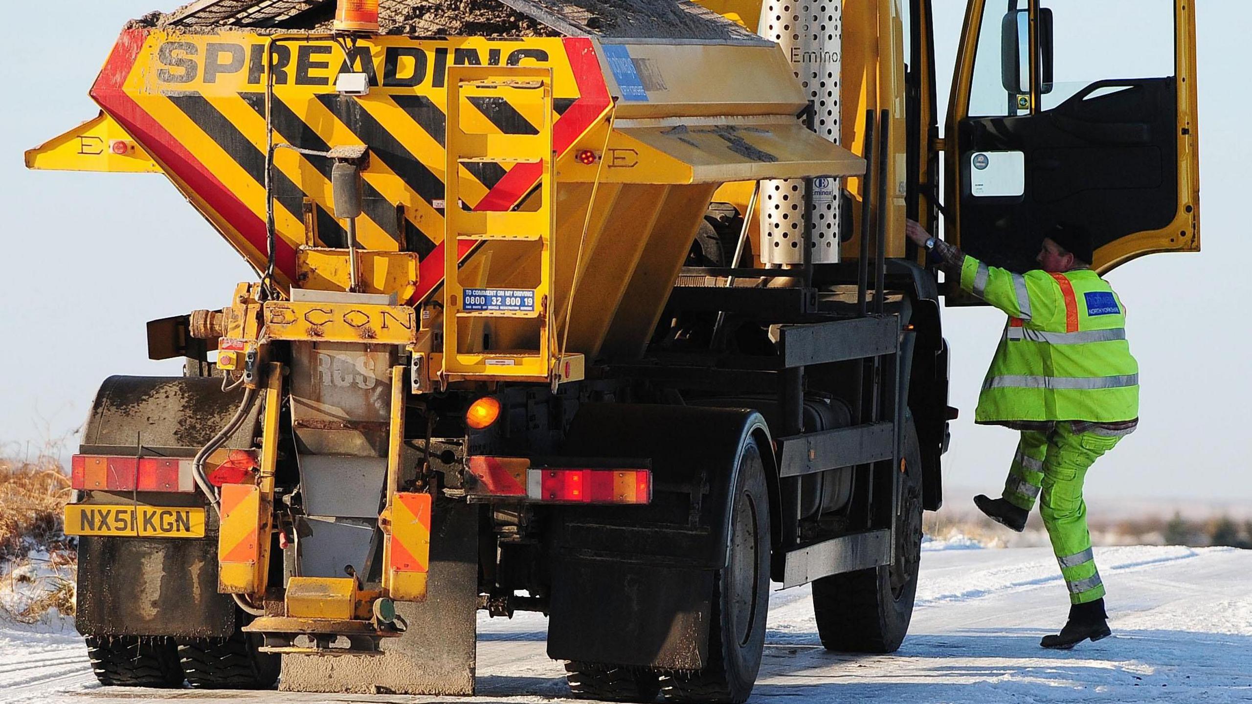 Gritting lorry parked on a snow-covered road, with a driver about to climb up to the vehicle's cab