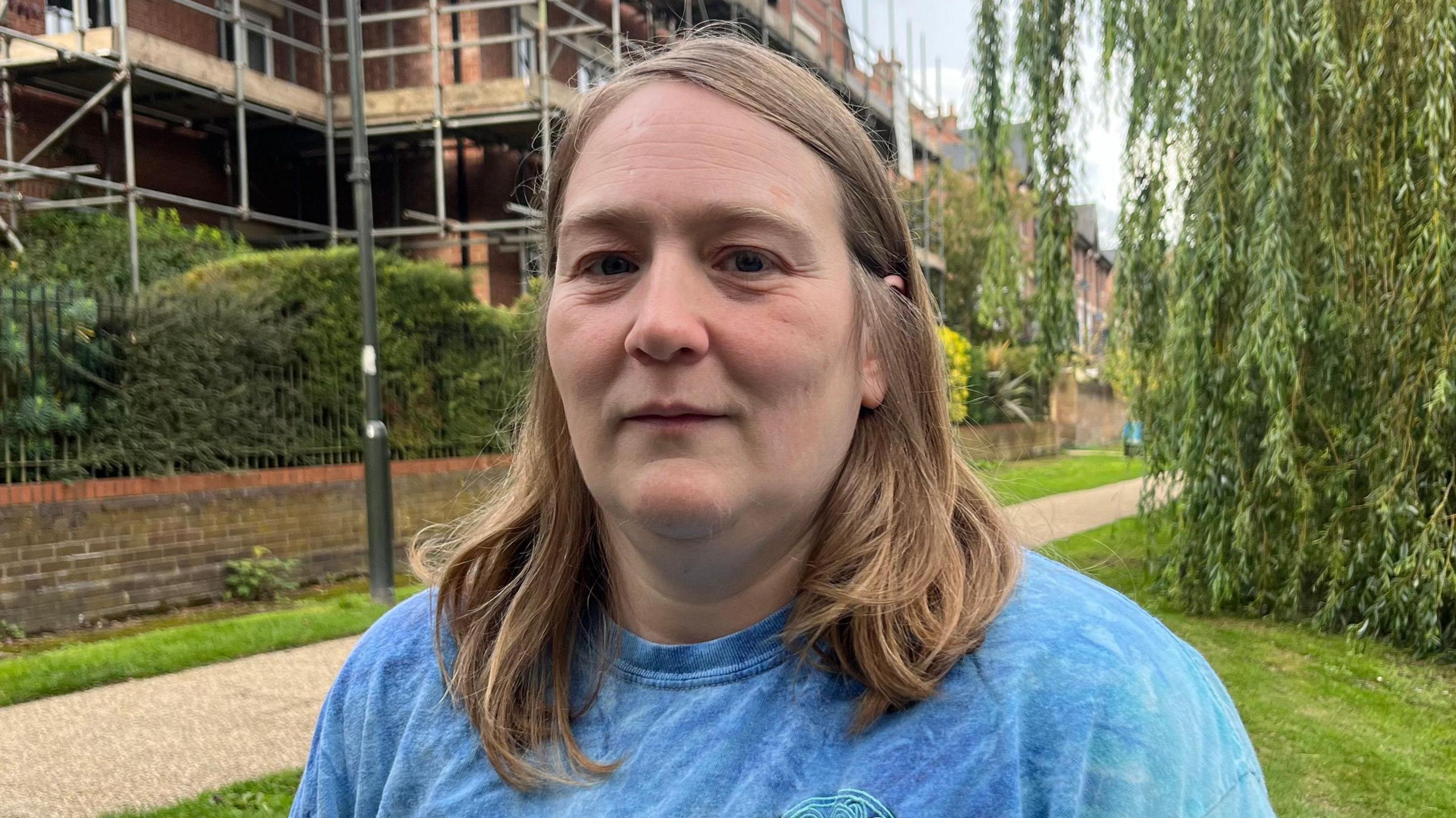 A close-up of a woman, Dr Eleanor Blakelock, wearing a blue top. She has shoulder-length light brown hair. She is standing in front of a building an there is a path and greenery.