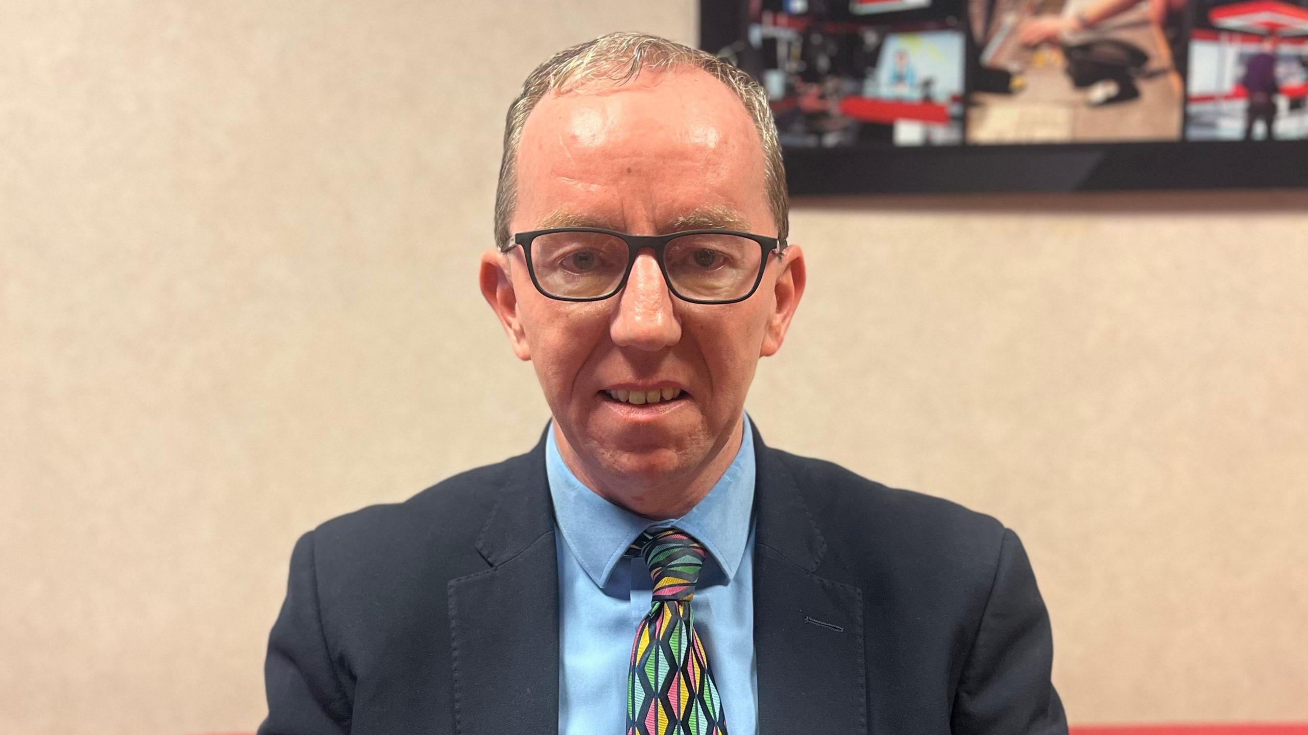 A man wearing a navy blazer with a light blue shirt and a blue, red and yellow tie that has diamond shapes on it. He has light hair and is wearing glasses. He is sitting on a red sofa.