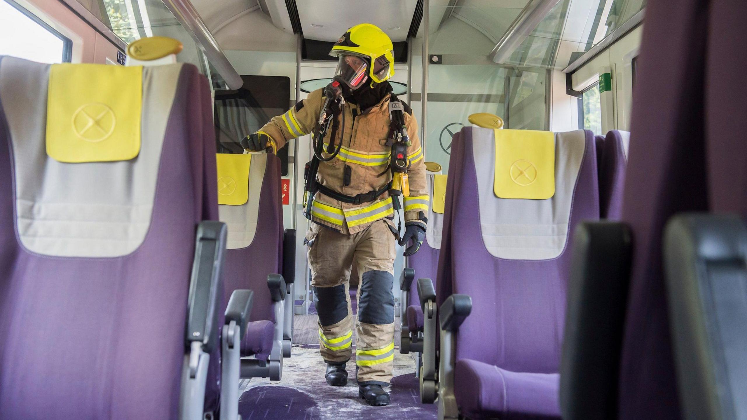 A firefighter wearing breathing apparatus walks through a train carriage containing purple seats.