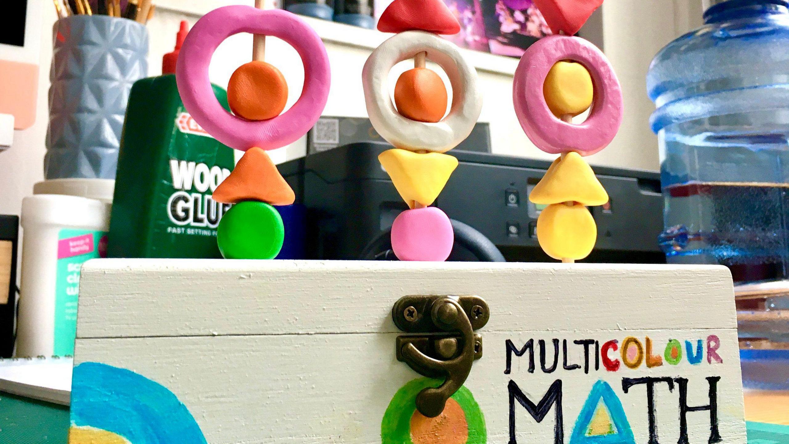 An image of a few coloured shapes made out of plasticine stacked on top of each other on a wooden dowel on a box with the words multicolour maths painted on to it on a desk