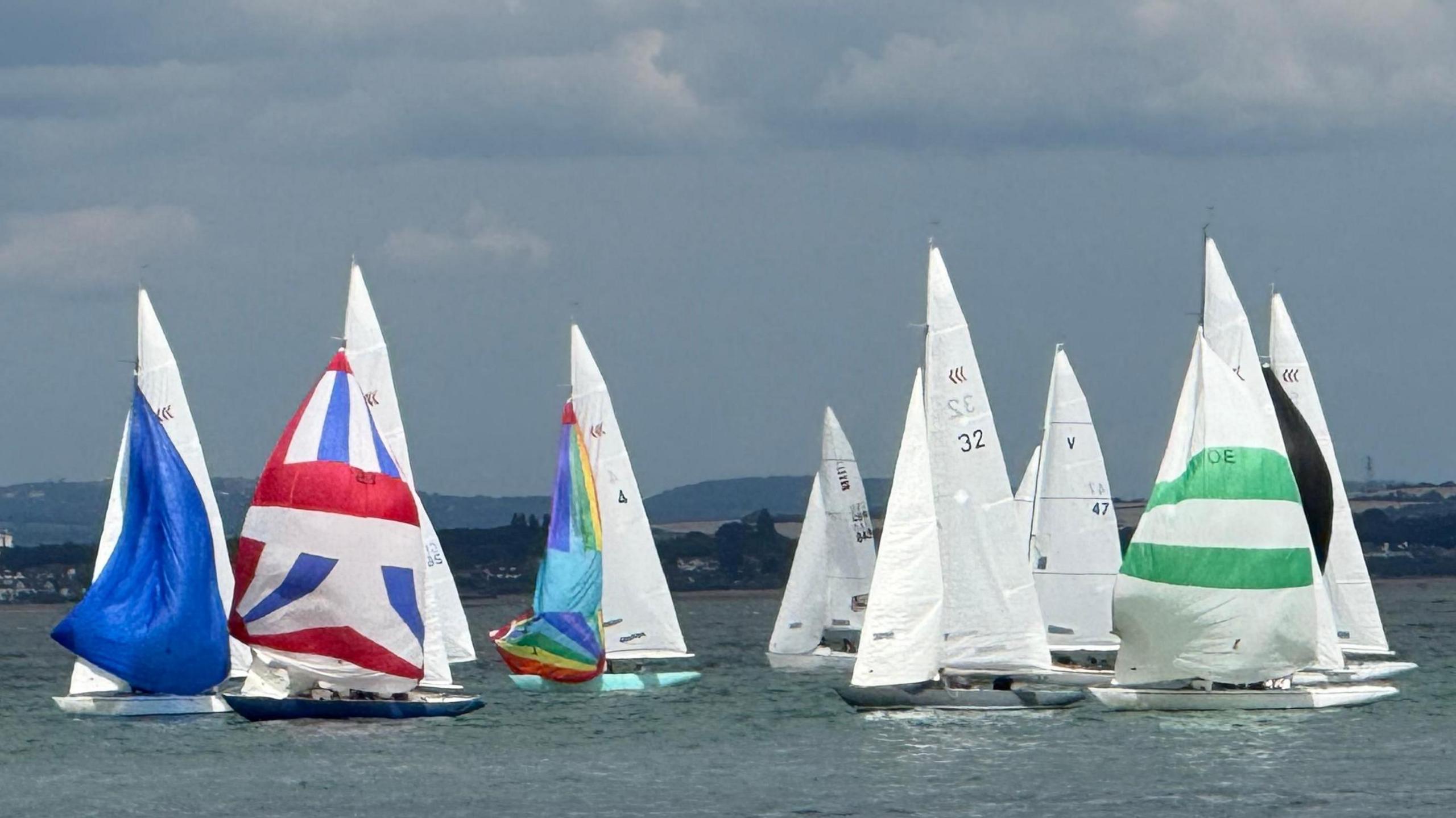 Several yachts battle it out on the sea in a race. Their sails are mostly white but some have multi-coloured spinnakers. The skies are grey with a hint of white clouds while the sun appears to be shining
