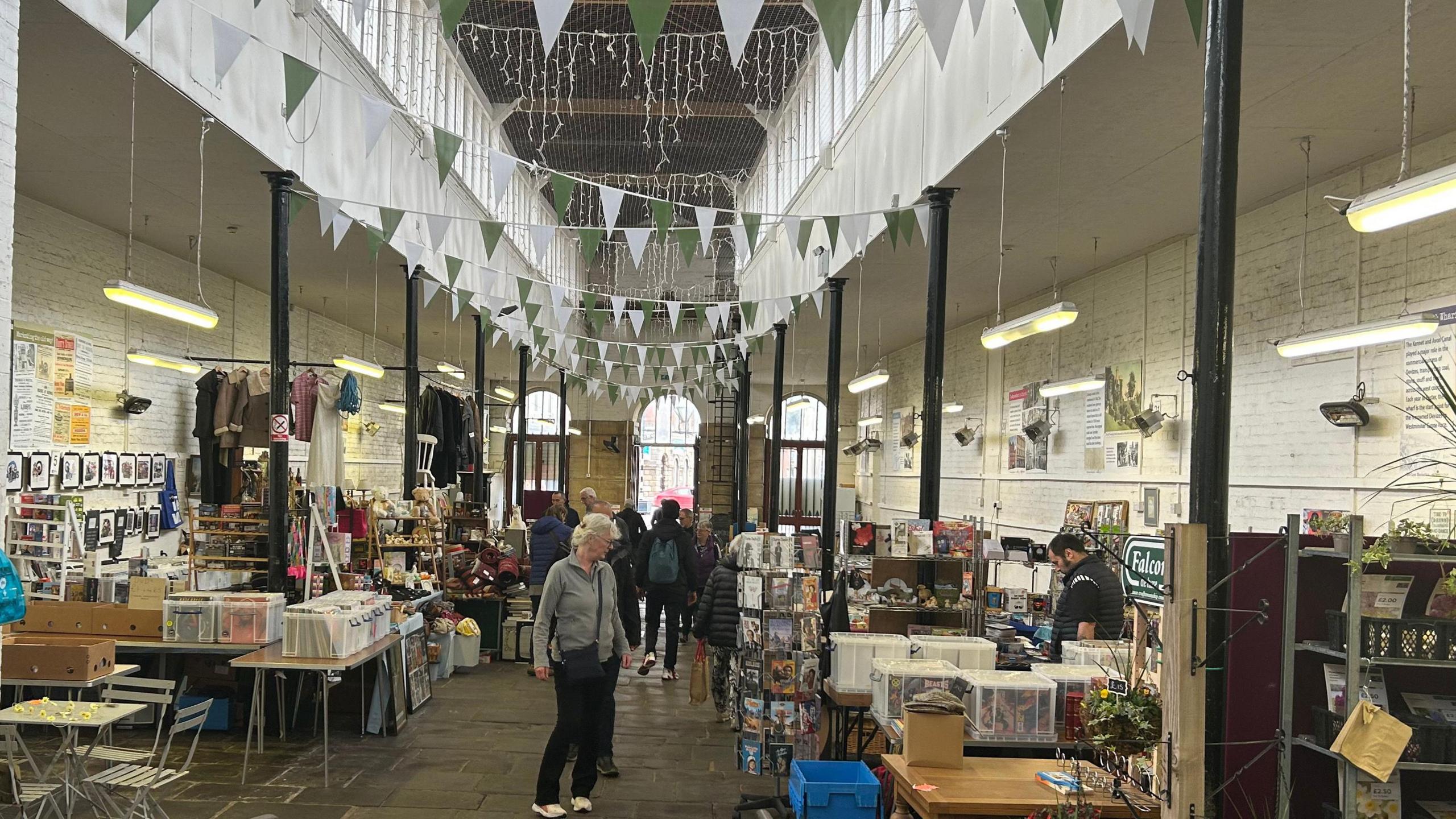 Inside the undercover market, a large room with flagstone floor, pillars down the sides, busy stalls with items and the public walking down the middle