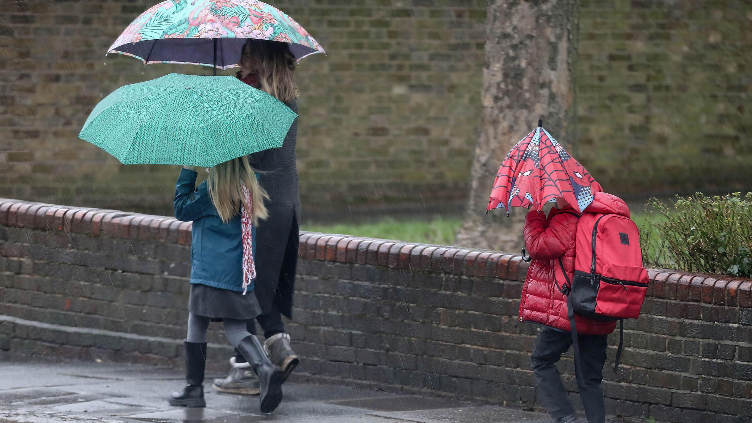 A woman walks with two children to school, all of them holding umbrellas. One is patterned with Spiderman pictures and is held by a child in a red puffer jacket and matching backpack. A girl carries a turquoise and black patterned brolly and wears black wellington boots. 