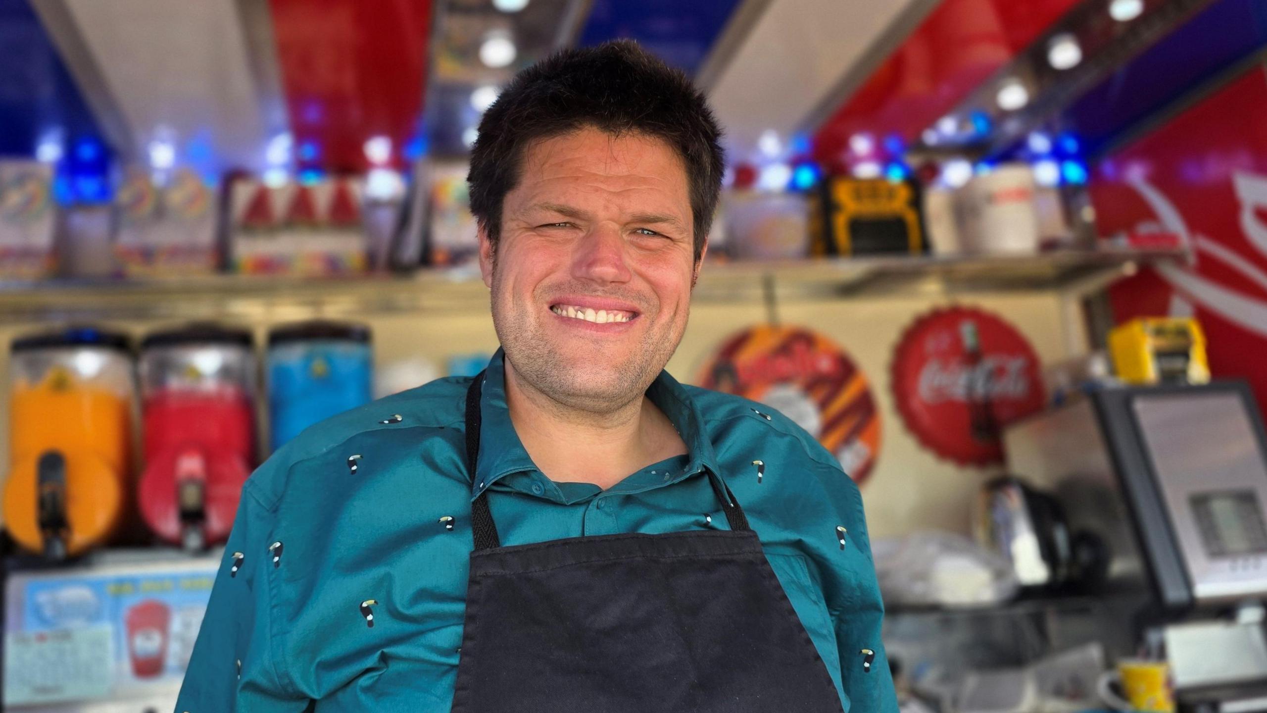Edward Scotter is standing in his kiosk. He has a dark apron over a blue/green shirt.