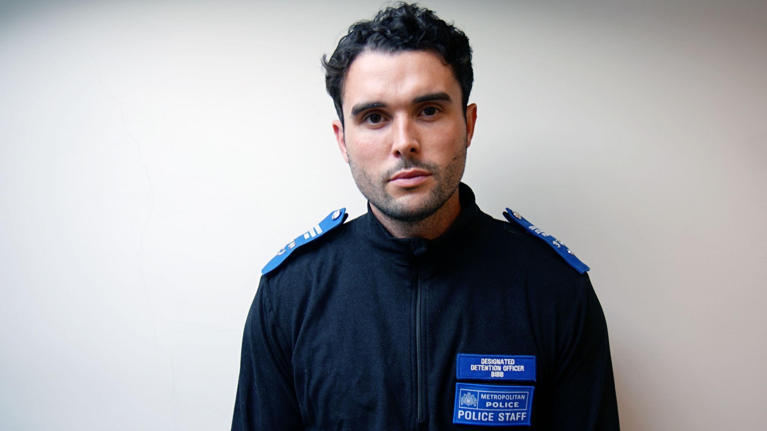 Rory Bibb, a young white man with dark, curly hair, dressed in the uniform of Metropolitan Police civilian staff, a navy zip-up top with a badge saying "designated detention officer" and royal blue epaulettes. He is standing against a blank white background.