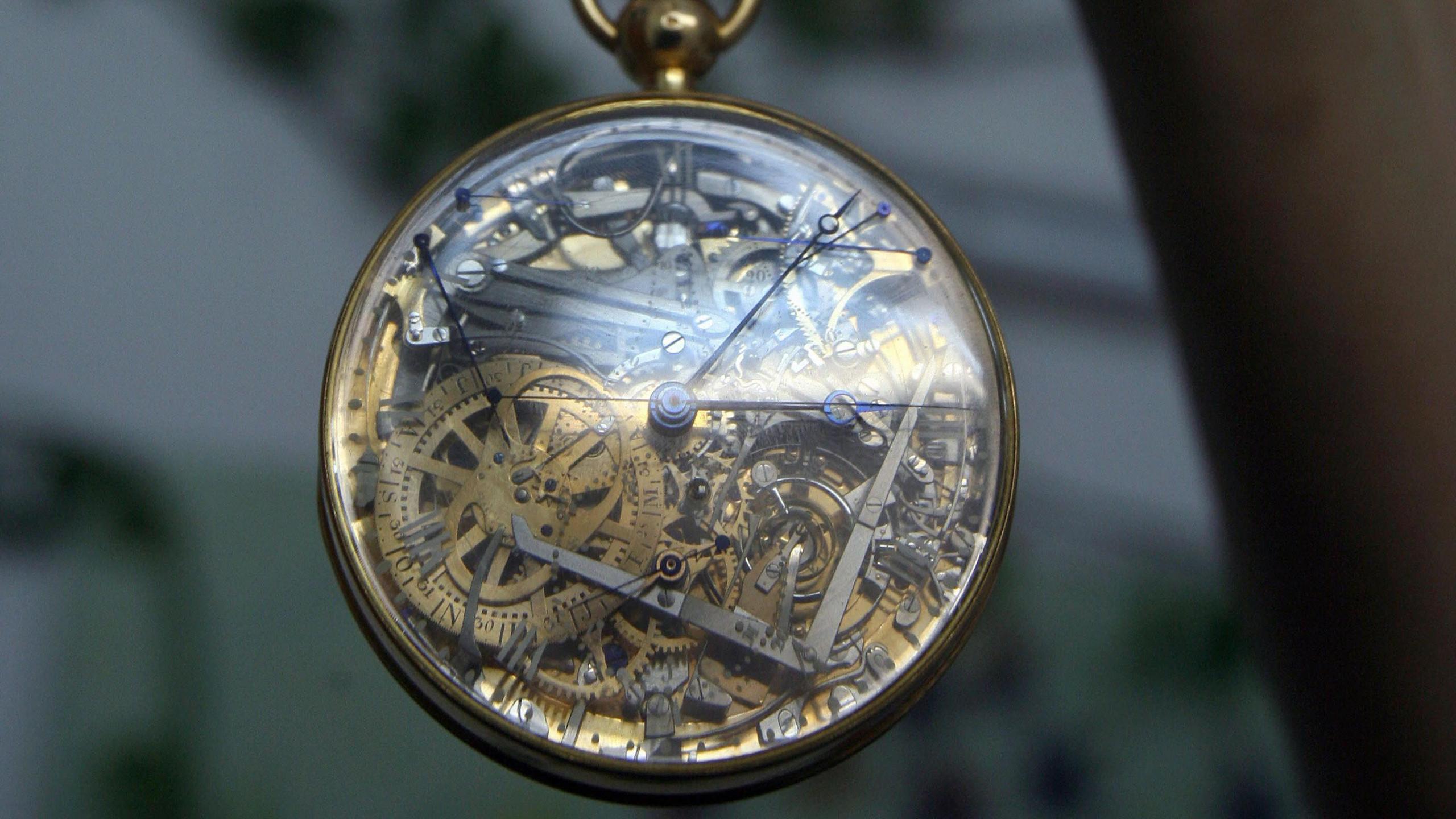 The gold casing of the watch with white roman numerals and a transparent crystal dial, showing hundreds of tiny platinum and gold parts underneath 