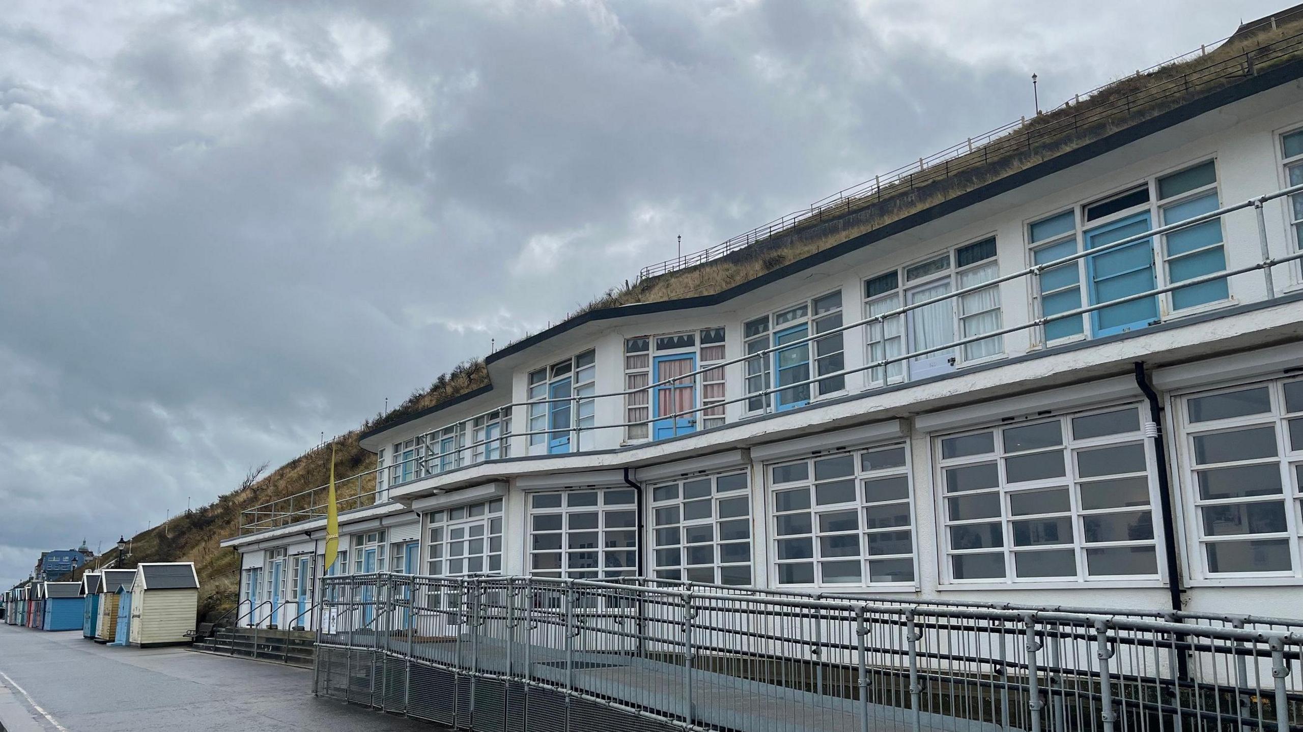 The seafront Artspace building: a 1930s-style white building with hundreds of panel windows and a few blue doors. It sits below a grassy cliff on a promenade, next to a series of pastel painted beach huts that recede into the distance. The sky is cloudy and grey.