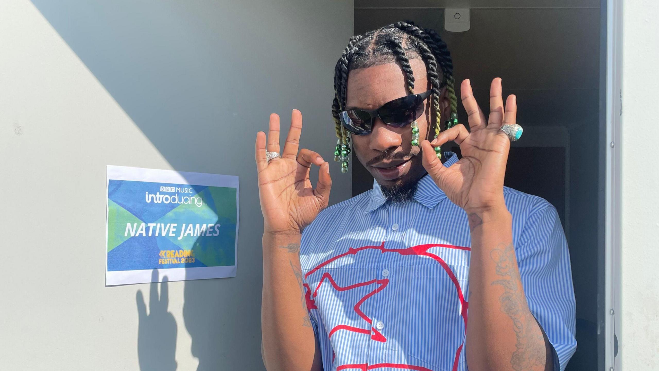 A man smiles at the camera while giving the 'OK' sign with both his hands. He is wearing a blue and white striped shirt with a red design on it. He is wearing sunglasses and has dark coloured braids. 