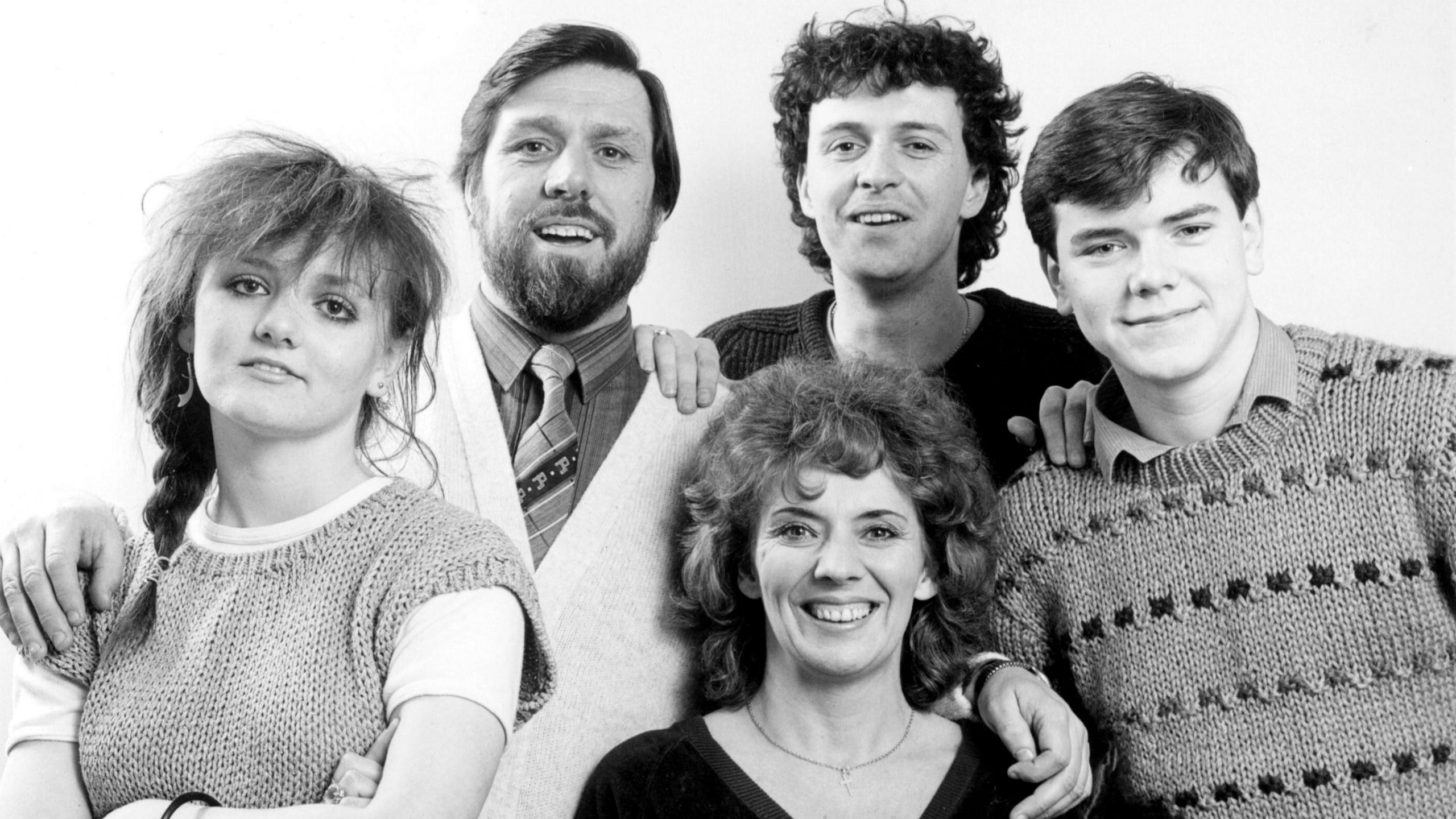 A black and white photograph of the Grant family, featuring parents Bobby, portrayed by Ricky Tomlinson, and Sheila Grant, played by Sue Johnston, along with their children Barry, Karen and Damon. They are all smiling at the camera and have their arms around each other.