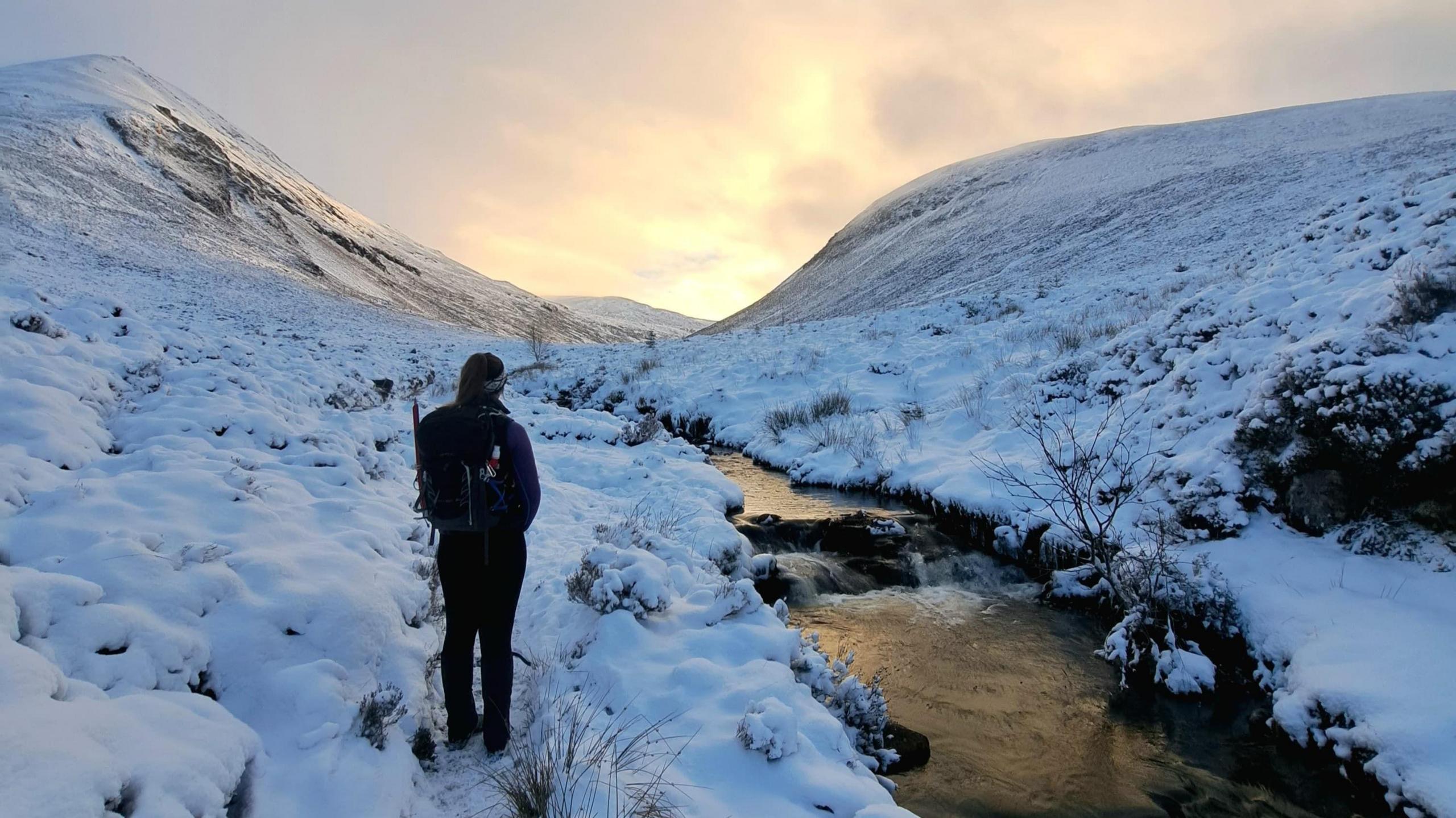 A woman stands with her back to the camera in a snowy landscape. a river runs down past her left hand side. In front of her is a dramatic mountain pass.