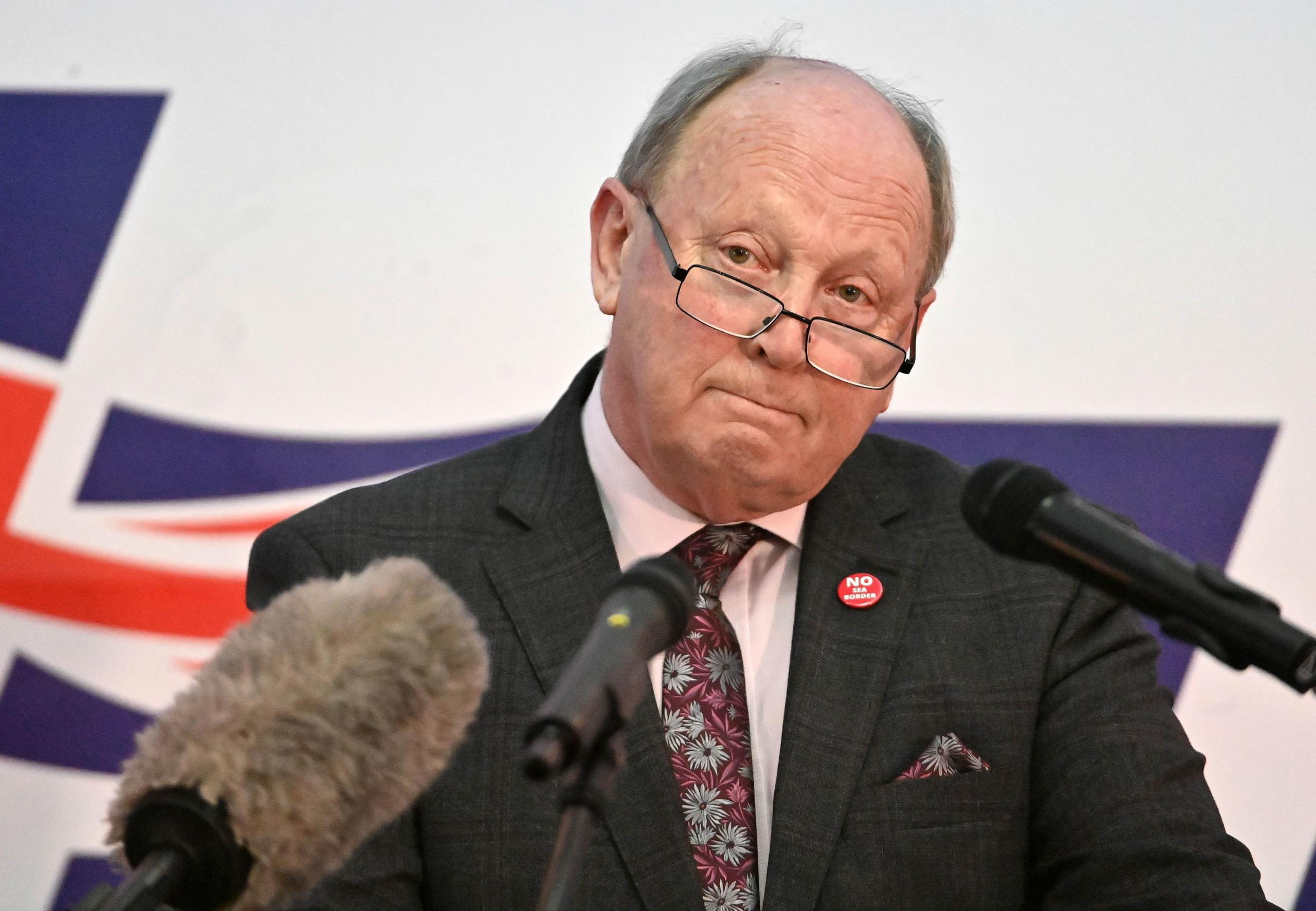 Jim Allister in black suit stood in front of microphones, union jacket visible in the background