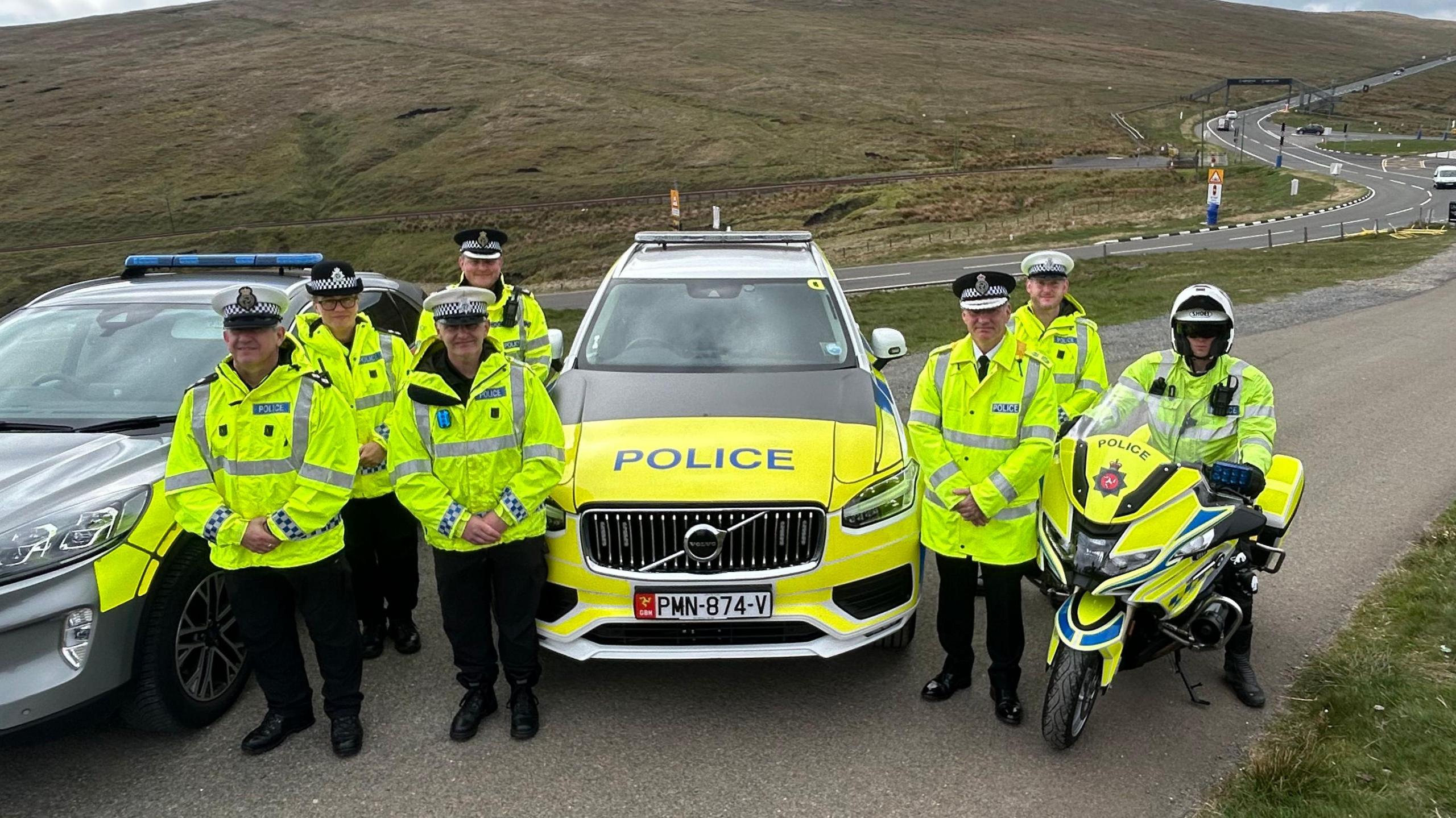 Seven police officers are positioned next to a police car, one is on a police bike. They are wearing hi-vis jackets and black trousers and boots and black and white hats. They stand next to the mountain road, part of the circuit TT riders race on during the festival.