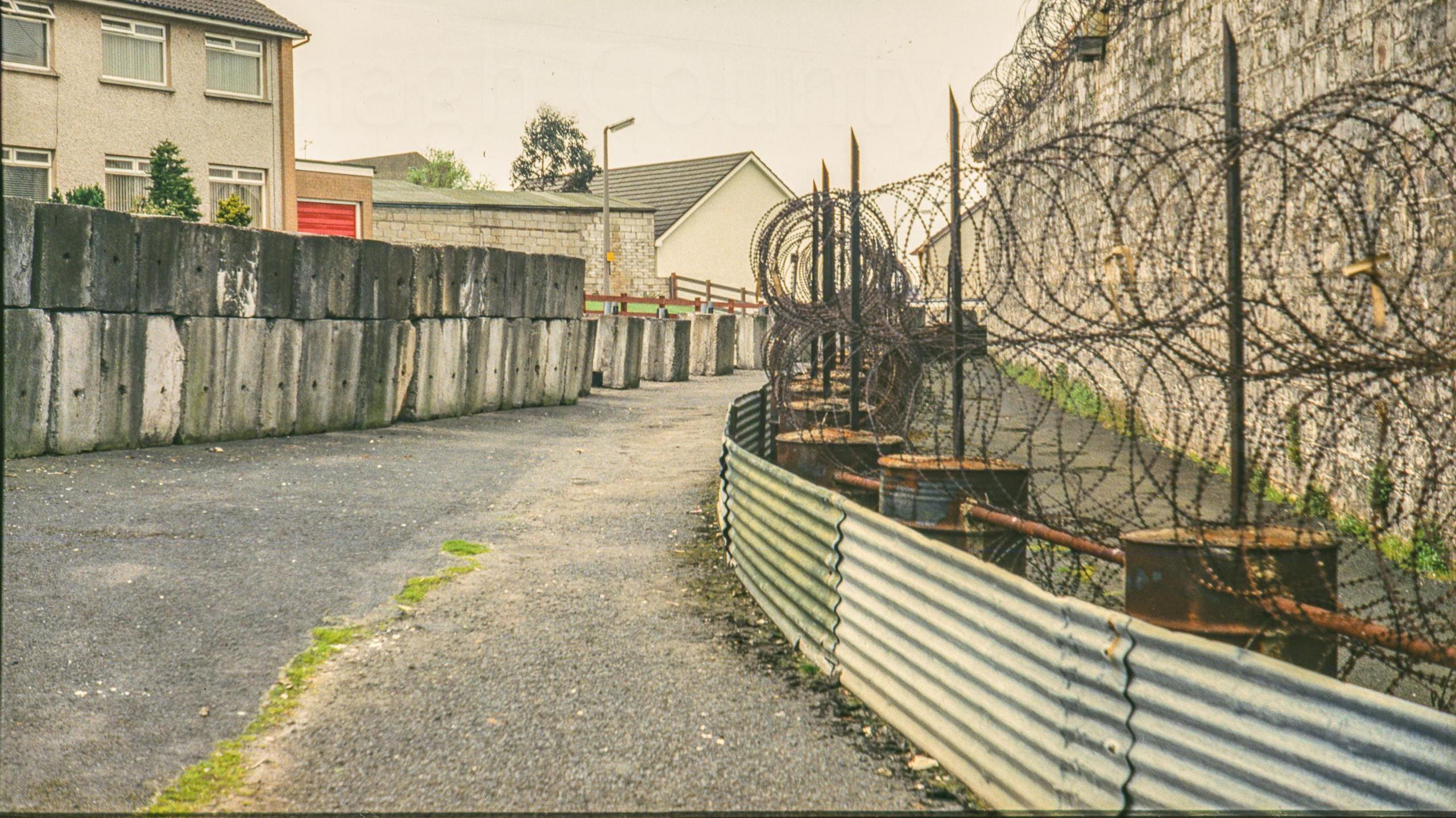 A digitalised photo from a film of a stone wall with barbed wire along the top.  Concrete bricks are across from the wall. Looking towards the Newry Road showing the prison wall fortified with large concrete blocks, sheets of corrugated aluminium and coils of barbed wire.