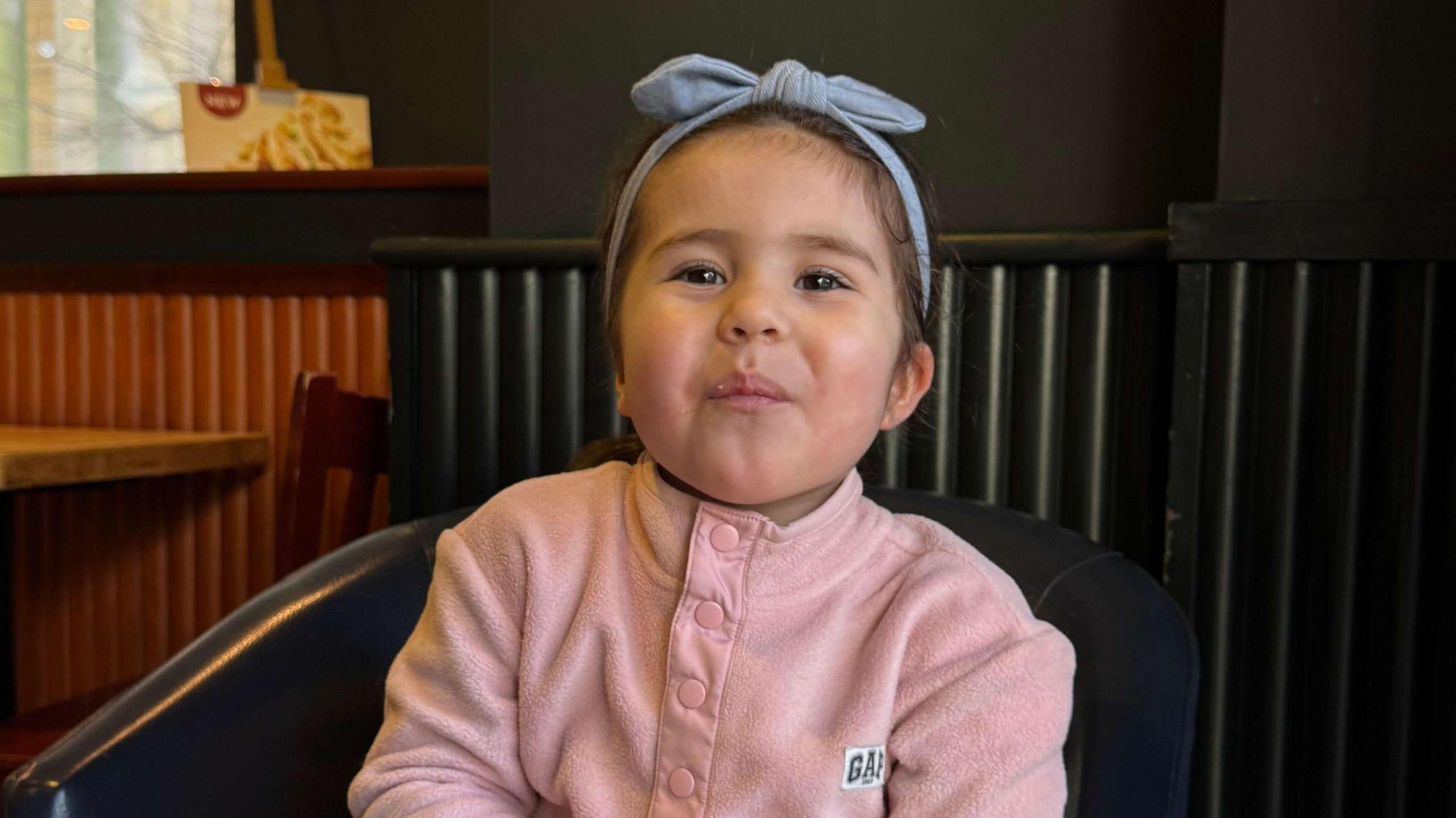 Louisa Palmisano smiles wearing a pink button-up jumper with a blue headband. She is sat in a chair next to a radiator indoors.